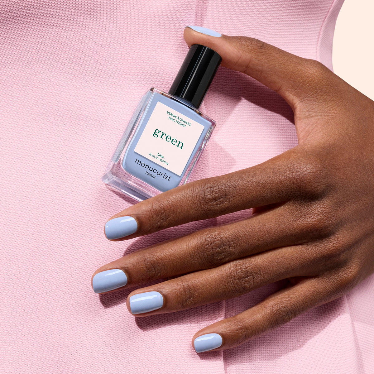 A hand with pastel blue Lilas nail polish holds a matching Manucurist bottle from the Green™ range against a pink fabric background.