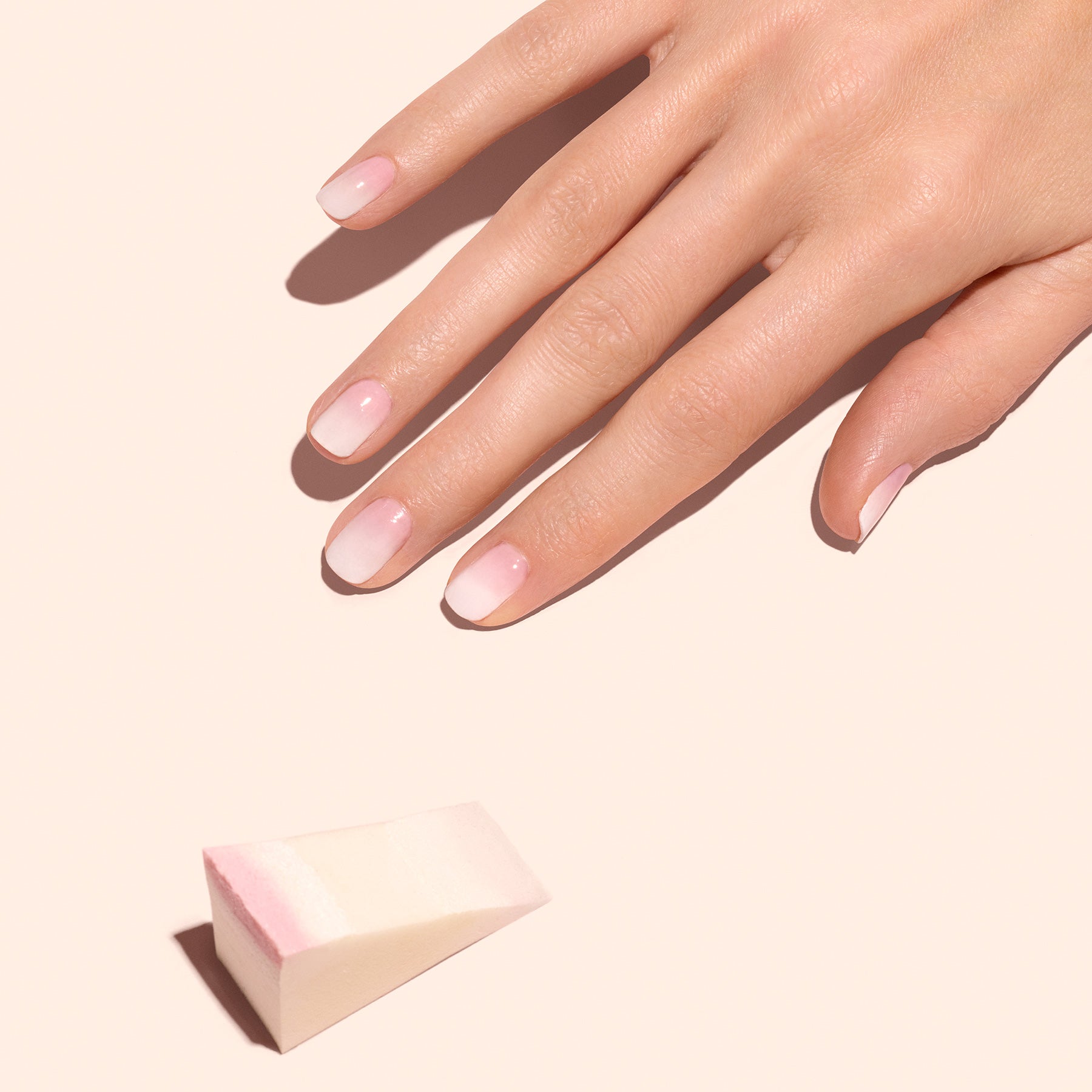 A hand with natural pink and white ombre nails rests on a peach background. Nearby are Manucurist’s Nail Art Sponges (x4), perfect for creating seamless aura nail gradients.