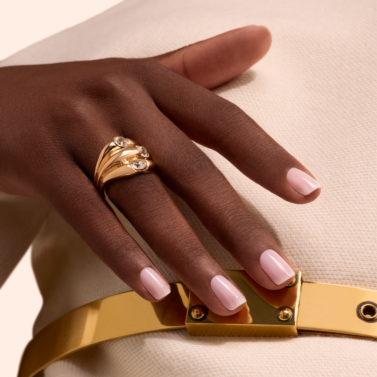 A hand with Manucurist Milky Pink gel polish nails wears a gold ring with three diamonds, resting on a white fabric outfit with a shiny gold belt.
