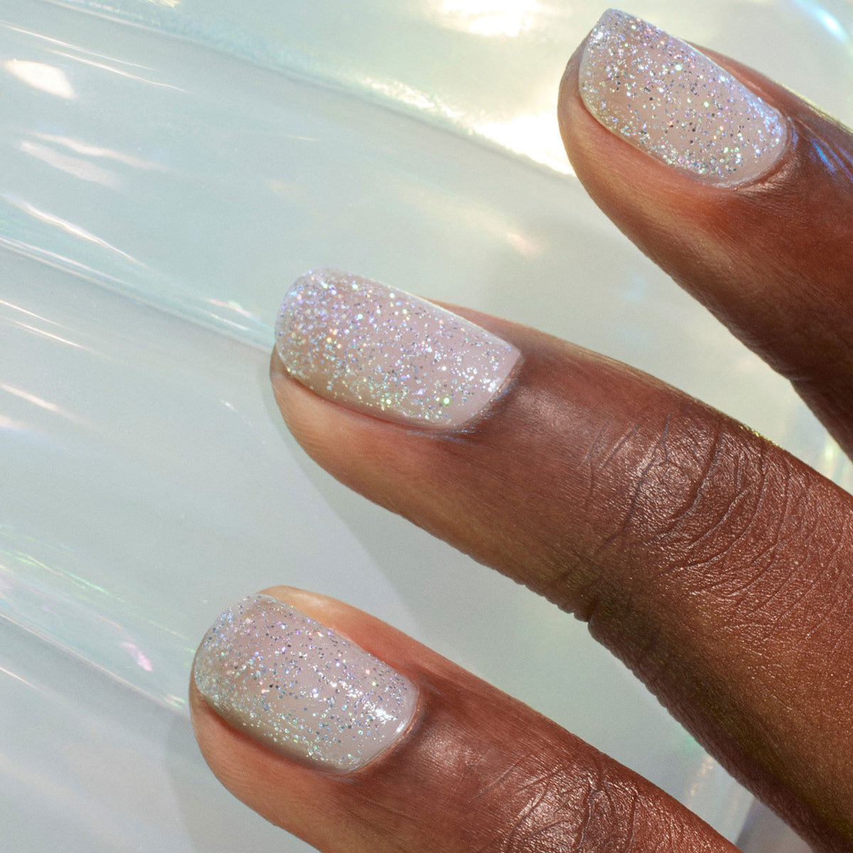 A close-up of three dark-skinned fingers shows nails painted with Midnight Swim, a sheer, glittery pale polish from Manucurist. The smooth, iridescent white background highlights this subtle nail art at its finest.