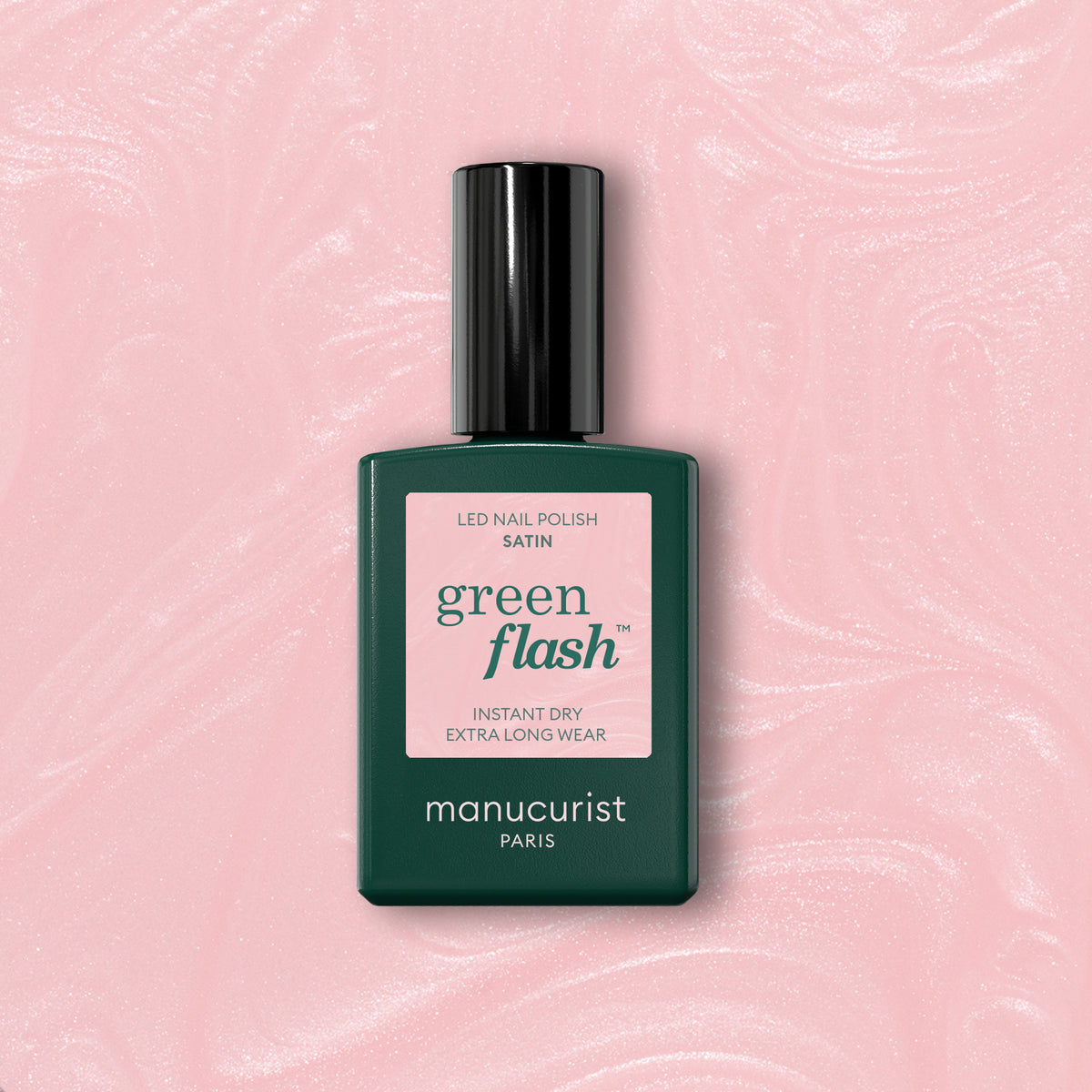 A bottle of Manucurist Satin Green Flash gel polish with a satin finish, in a dark green rectangular bottle with a pink label, is placed on a shimmery light pink textured background.