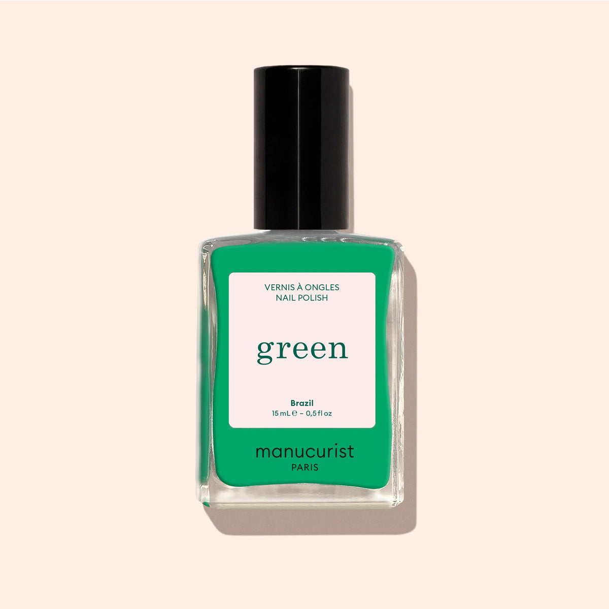 A bottle of Manucurist Brazil nail polish from the Green™ range, featuring intense electric green color and a black cap, is photographed against a pale beige background. The label shows green and pink colors with text.