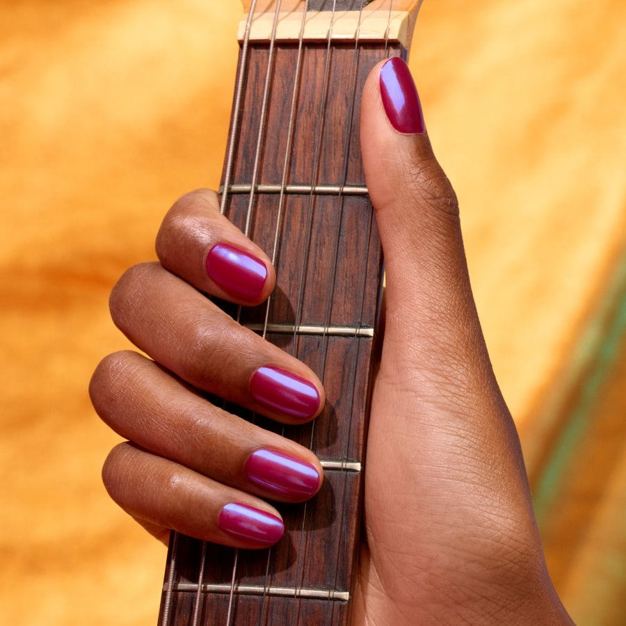 A hand with shiny magenta nails, highlighted by Manucurist Blue Glazed Powder’s pearly blue shimmer, holds a guitar neck against a blurred orange background.