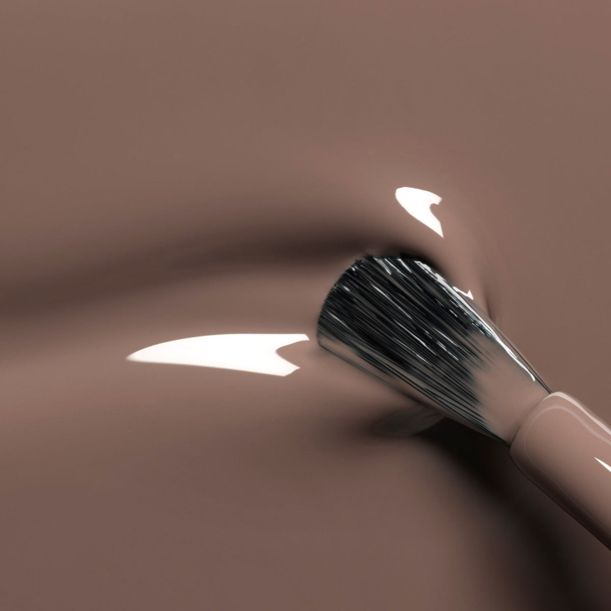 A close-up of a makeup brush applying Manucurist’s Freckles, a glossy brown nail polish with cool undertones, onto a smooth surface with reflections of light on the liquid.