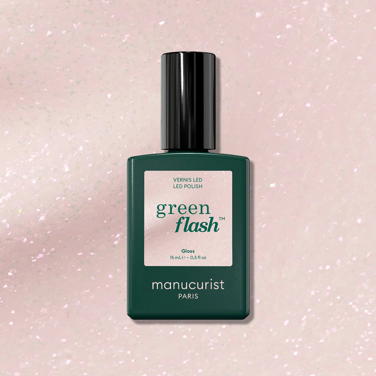 A green rectangular bottle of Manucurist Gloss sits on a light pink, glittery background, its label highlighting extra long wear and gloss that amplifies the iridescent pink shimmer around it.
