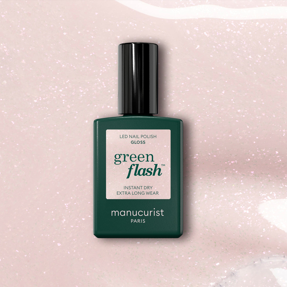 A green rectangular bottle of Manucurist Gloss sits on a light pink, glittery background, its label highlighting extra long wear and gloss that amplifies the iridescent pink shimmer around it.