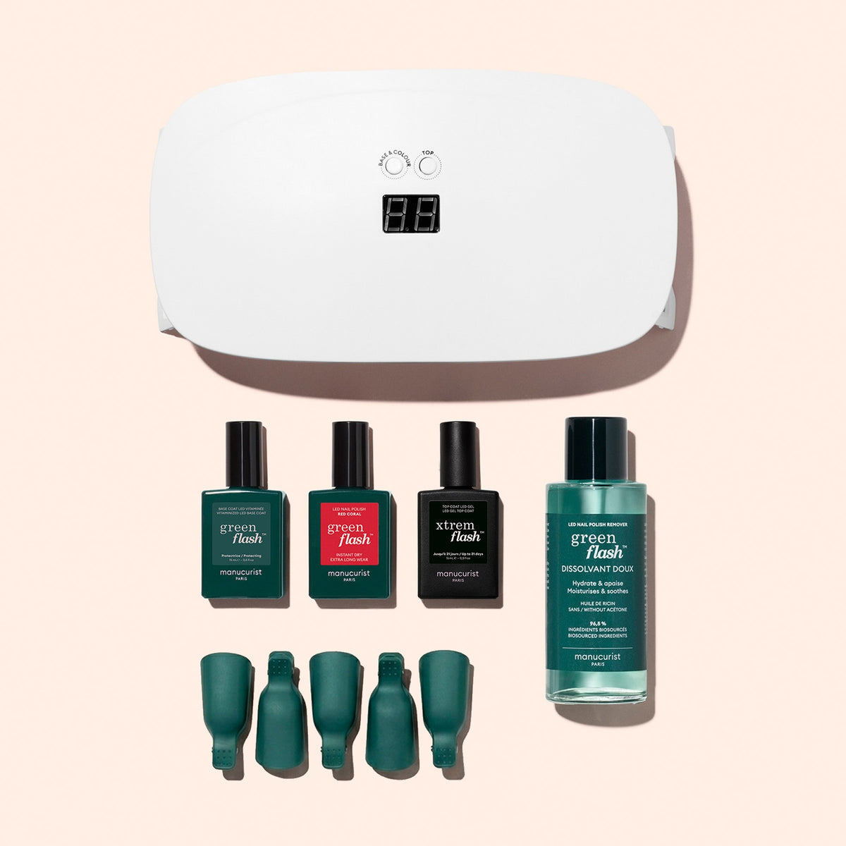 The Manucurist Discovery Kit, featuring a white nail lamp, three bottles of long-lasting nail polish, nail polish remover, and four green nail clips, is neatly arranged on a light pink background.