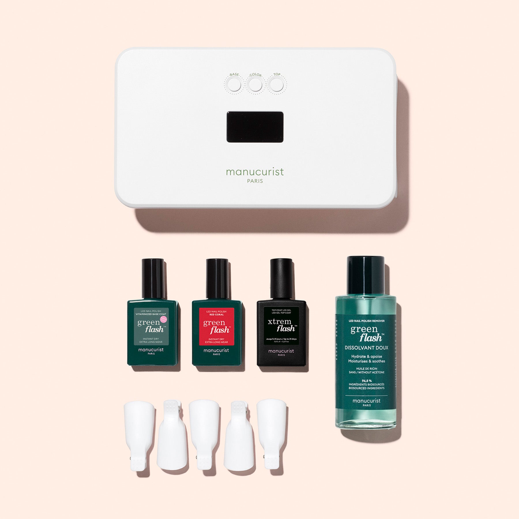 The Manucurist Discovery Kit includes a white LED lamp, three green nail polishes, a vitaminized base coat, a green remover bottle, and four white nail clips—perfectly arranged on a pink background for long-lasting nails.
