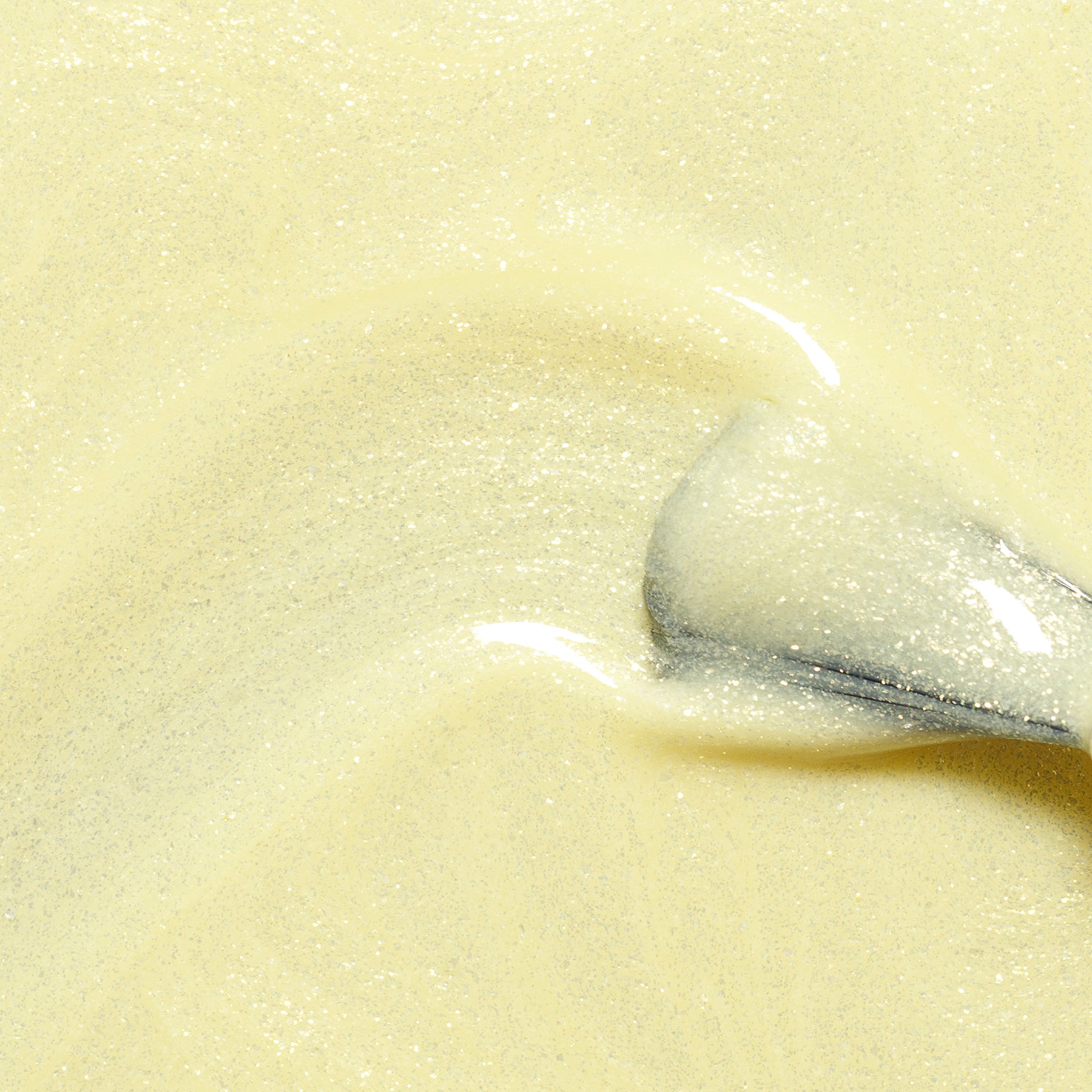 A close-up of a metal spoon stirring thick, creamy Lemonade by Manucurist, featuring pale yellow swirls and glittering highlights in the smooth texture.