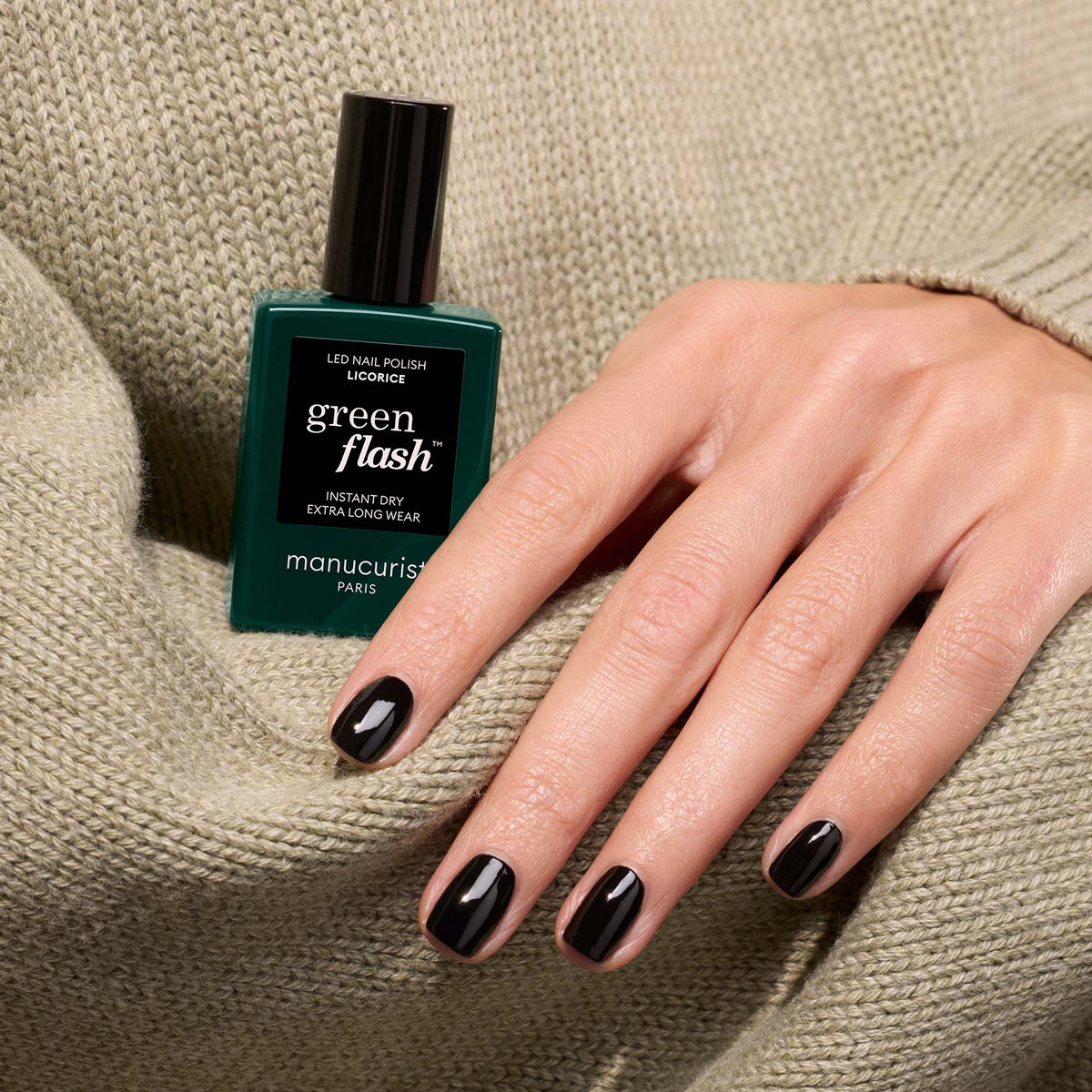 A hand with neat black gel-manicured nails rests on beige fabric beside a bottle of Manucurist Licorice polish.
