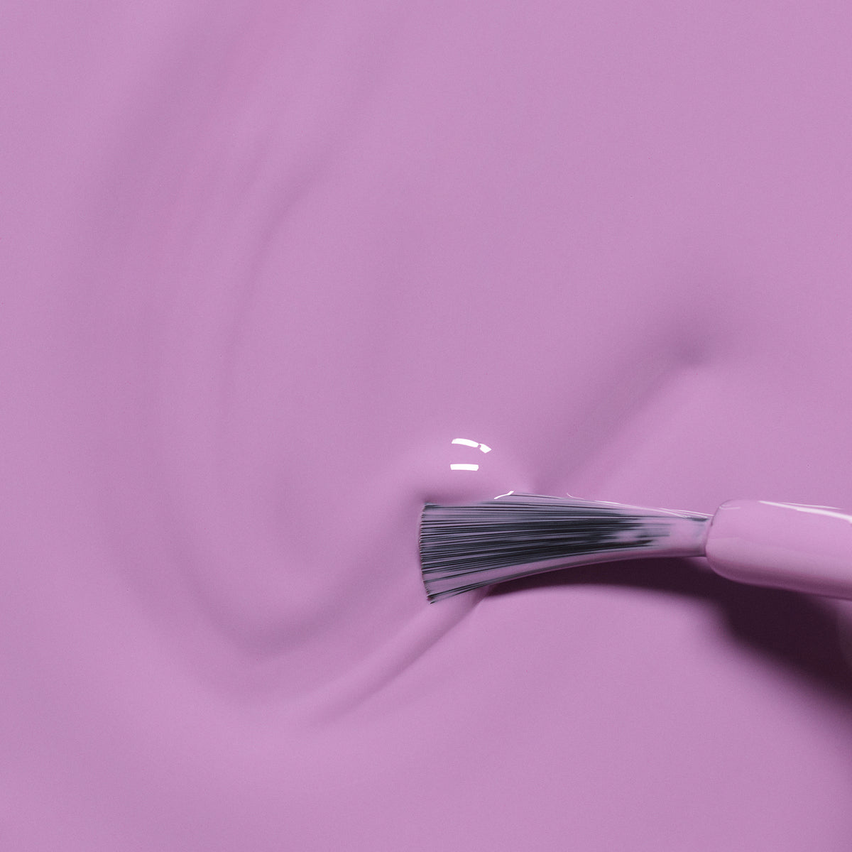 A close-up of a brush spreading Manucurist’s Lily the Unicorn, a vibrant, non-toxic pink and purple nail polish, in smooth, wavy strokes across a flat surface.