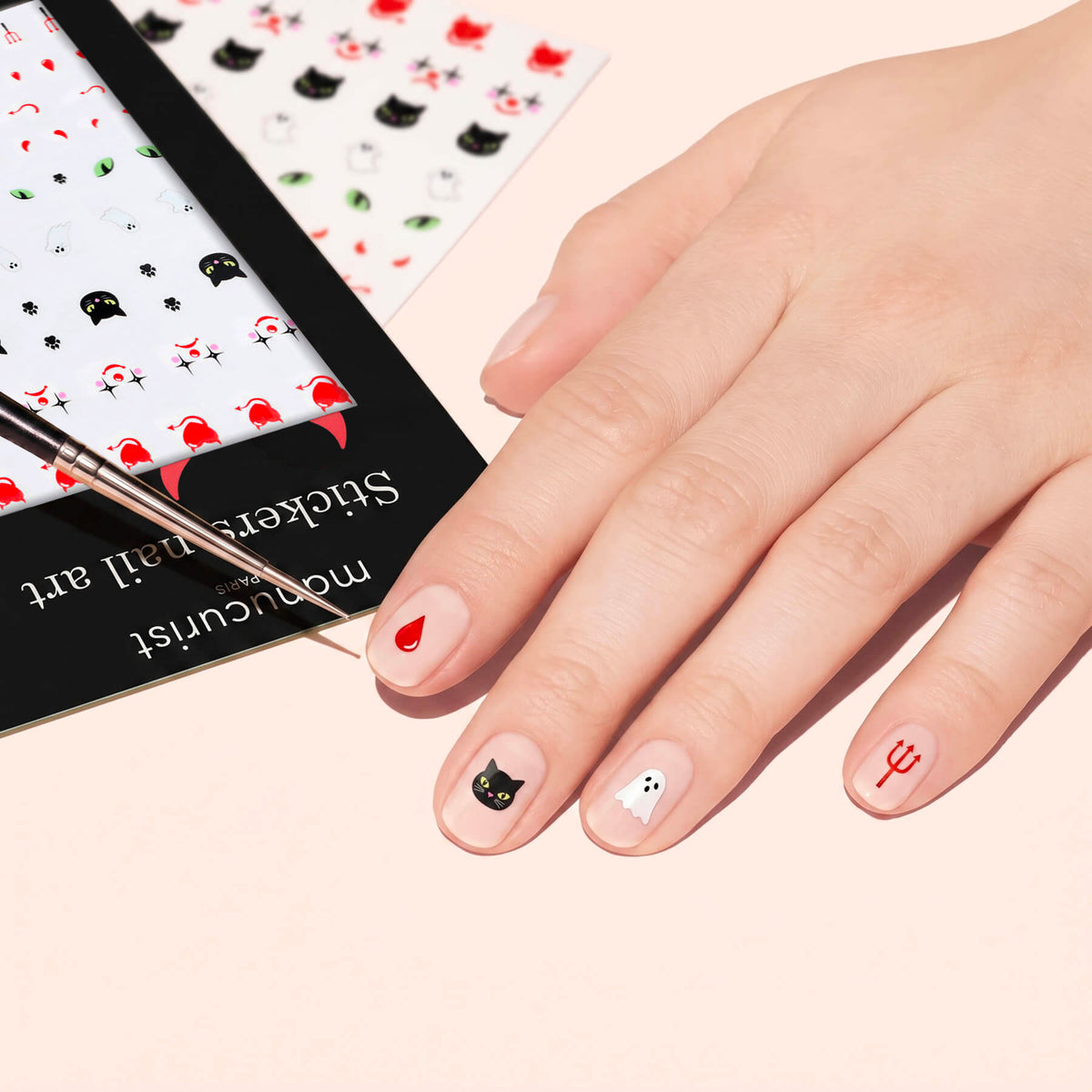 A hand shows off Manucurist Halloween Nail Art Stickers—cat, ghost, pitchfork blood drop—on nude nails. A sticker sheet and brush are nearby, great for festive Halloween manicures.