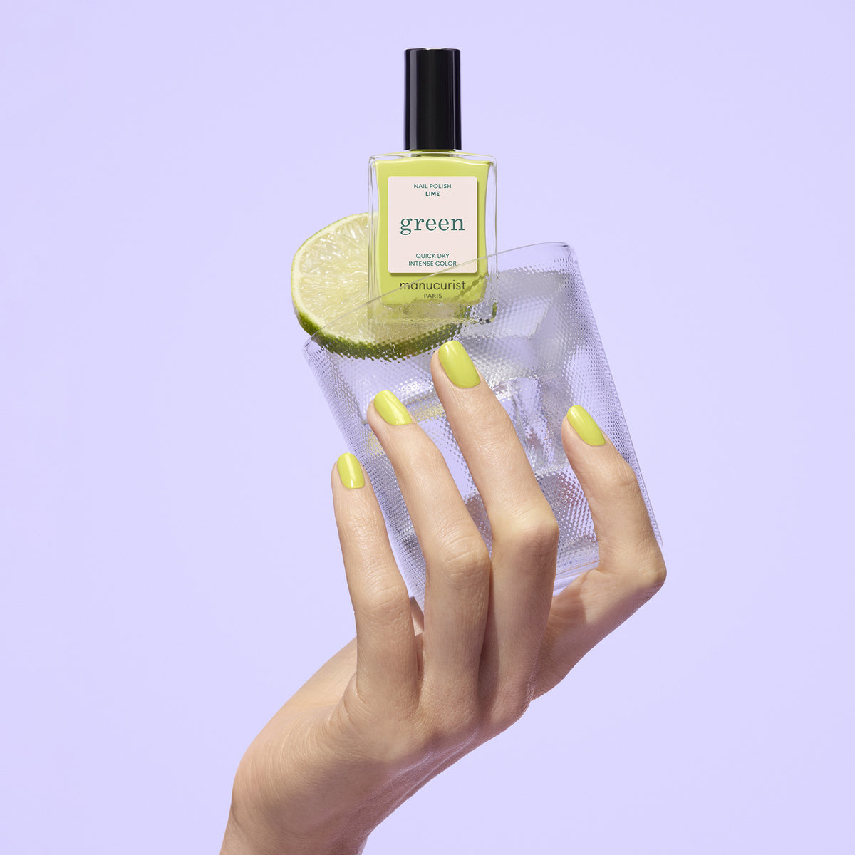 A hand with anise green manicured nails holds a textured glass with a citrus slice, as a Manucurist Lime polish bottle balances on top—all set against a light purple background.