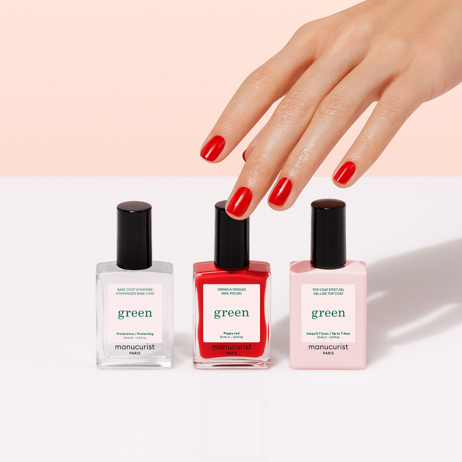 A hand with glossy red nails reaches toward Manucurist's Basic Kit—featuring three nail products in clear, red, and light pink—displayed on a white surface against a pale pink background.