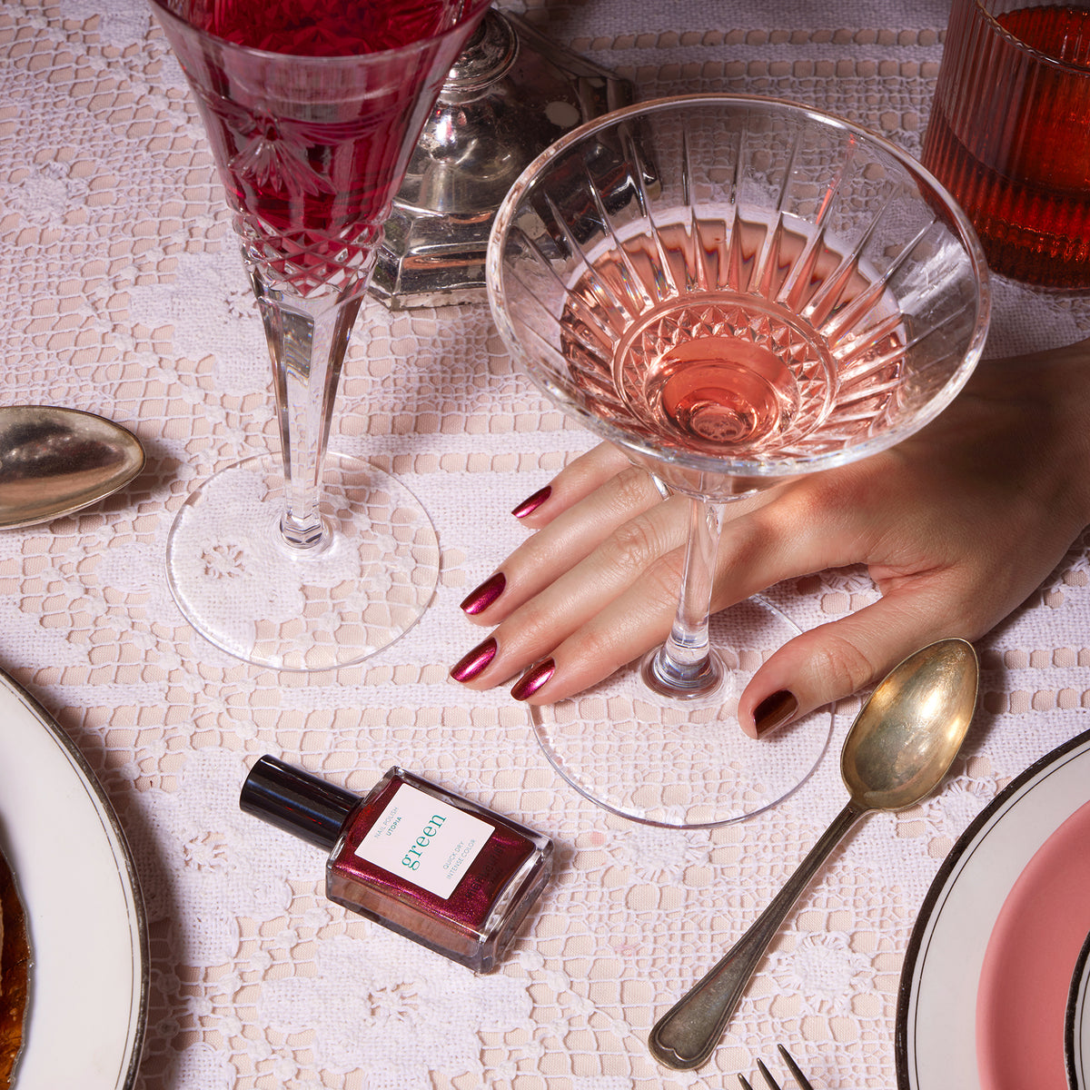 A hand with shimmering Manucurist Utopia nail polish holds a glass of pink wine on a lace tablecloth, accompanied by silverware, a plate, a wine glass, and a bottle of Green-labeled red nail polish.