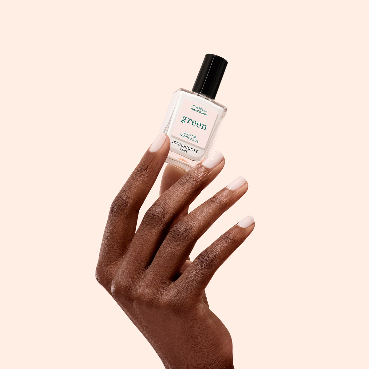 A hand with neatly manicured nails holds a clear bottle of Milky White nail polish by Manucurist against a plain light beige background.