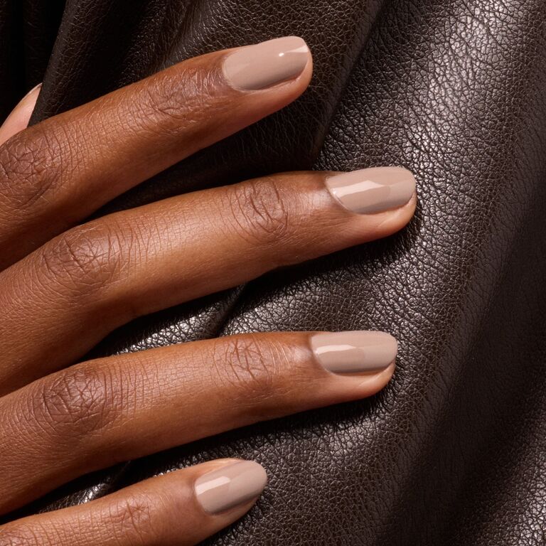 A hand with neatly manicured nails painted in glossy Dove Beige nail polish by Manucurist rests on textured dark brown leather fabric.