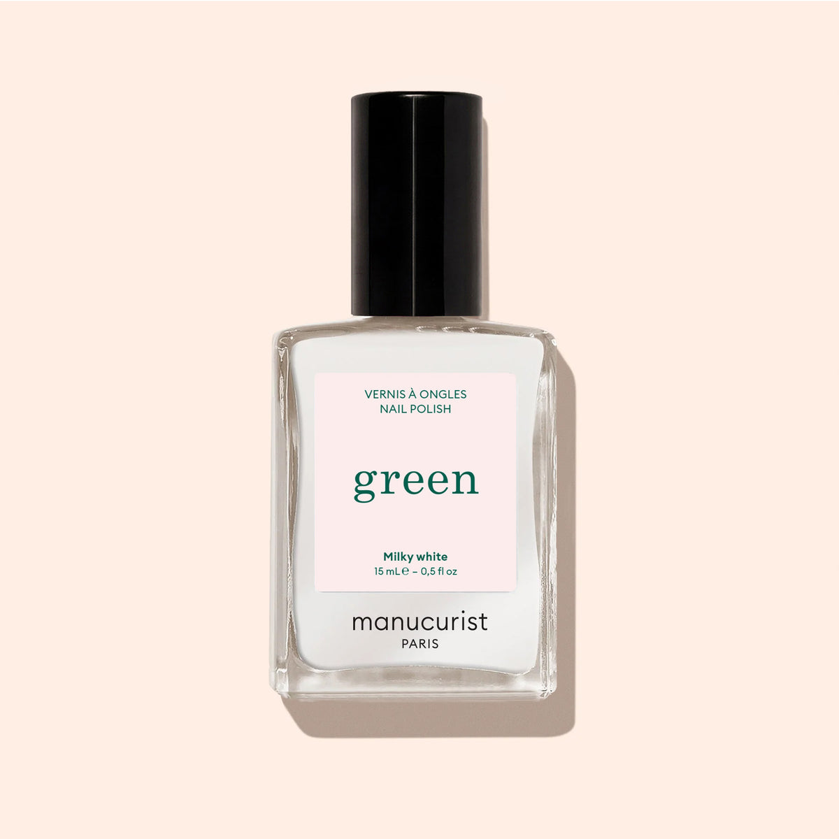 A clear glass bottle of Manucurist Milky White nail polish with a black cap and pink label sits against a soft beige background. The label reads: Milky White, green, Quick Dry Intense Color.