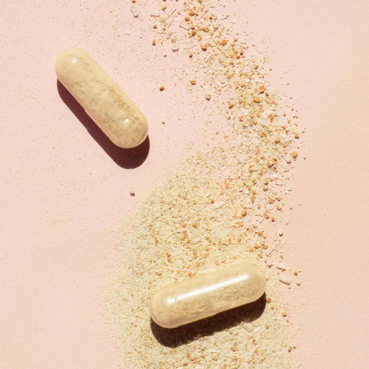 Two beige Manucurist Nail Boost capsules, formulated to strengthen nails, rest on a light pink surface with some of their powder contents spilled and scattered nearby.