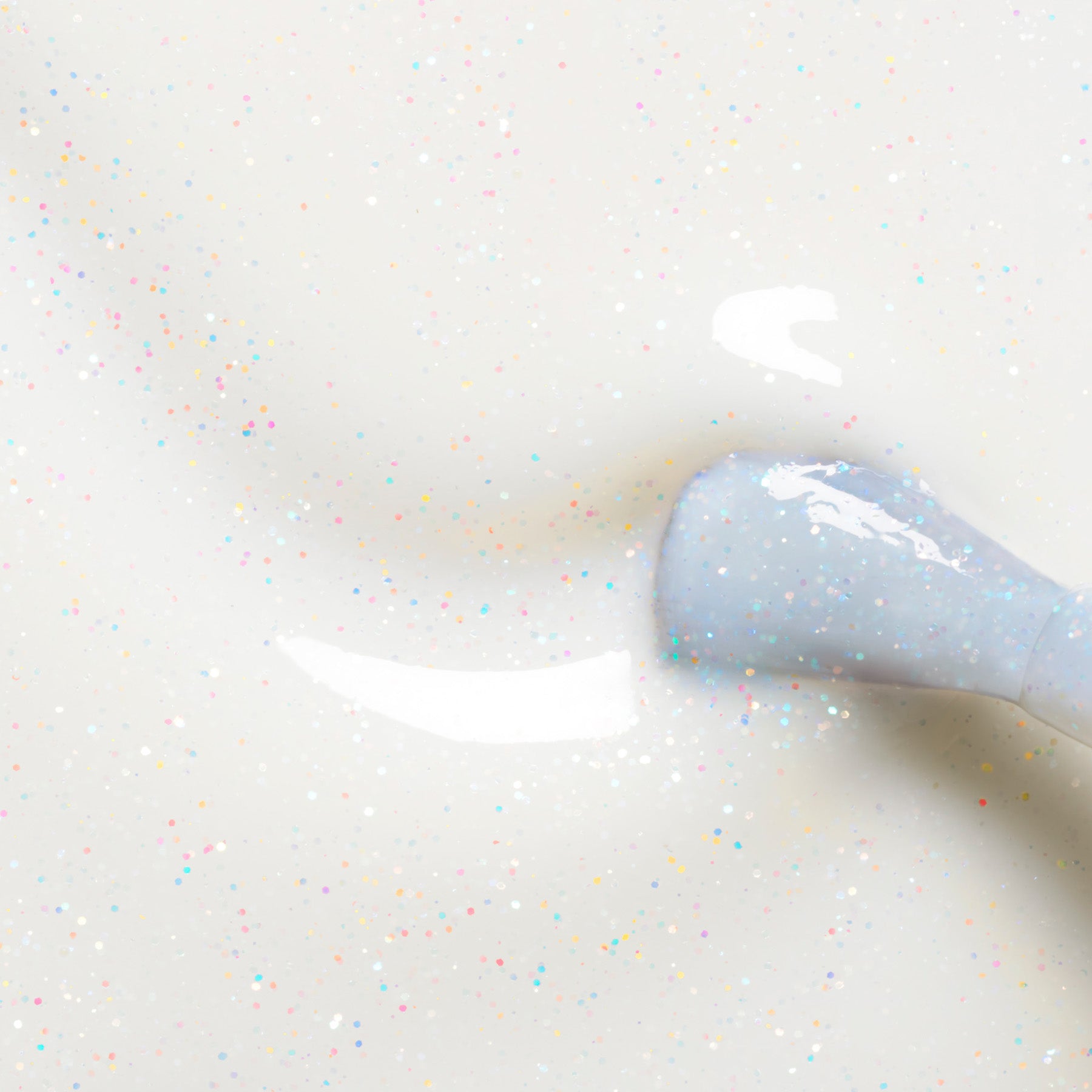 A close-up of a nail polish brush applying Nebula, a milky white polish with tiny colorful glitter specks from Manucurist, on a smooth surface.