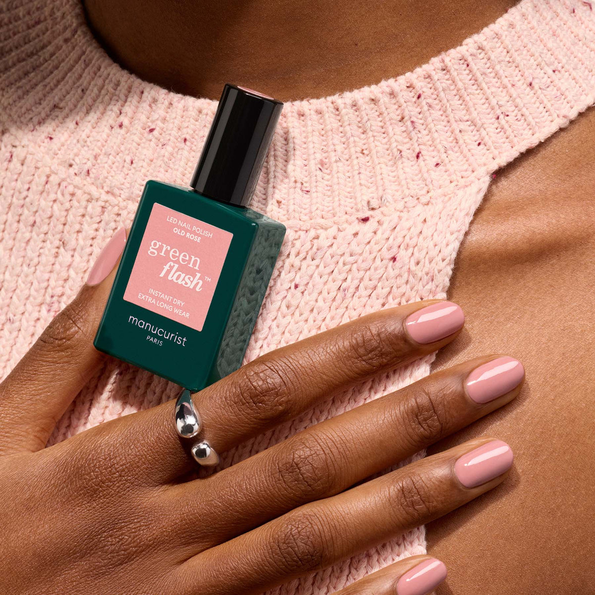A hand with Old Rose gel polish by Manucurist rests on a light pink knitted sweater, holding a green Manucurist bottle. A silver ring decorates the ring finger.