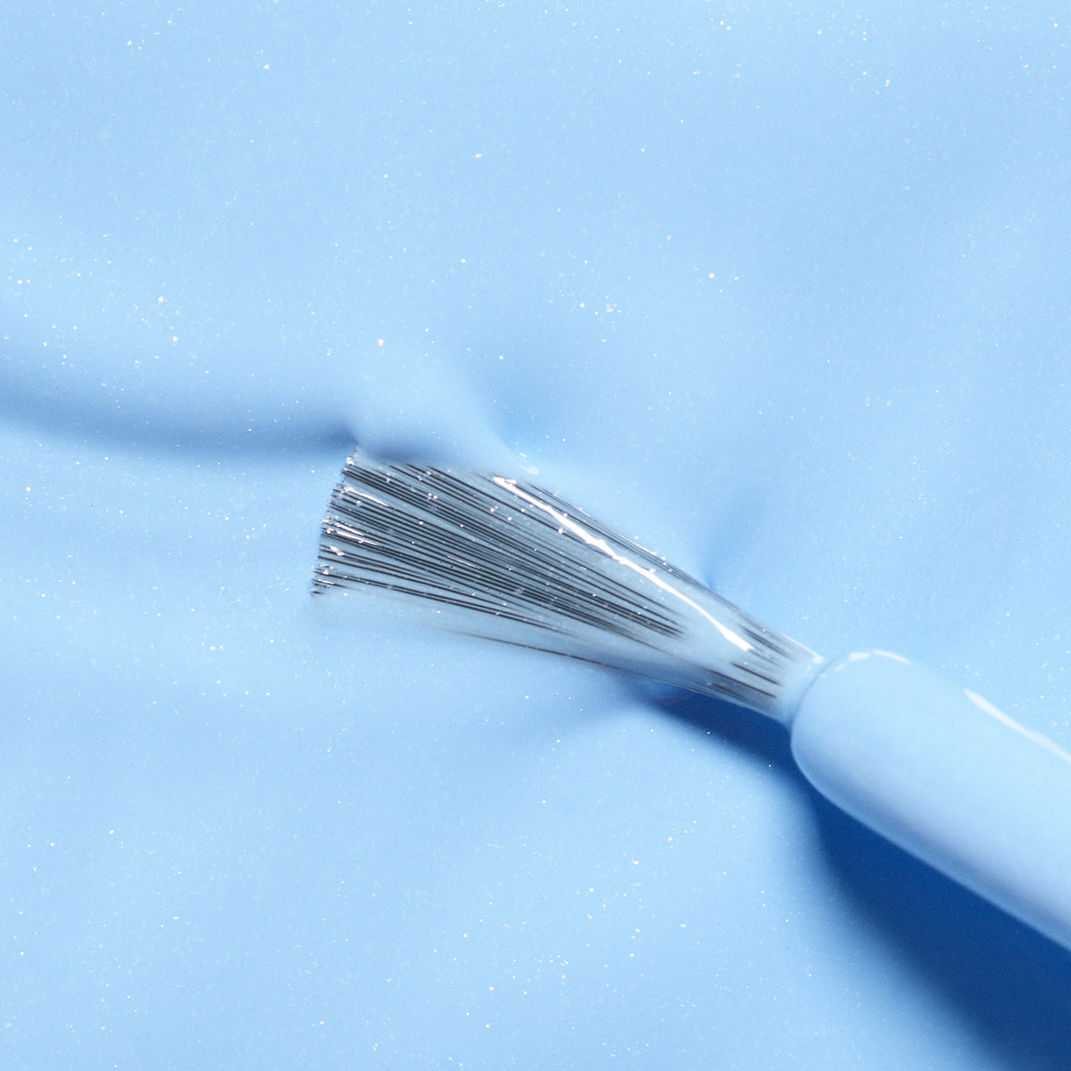 A close-up of Manucurist’s Pomme the Fish brush with silver bristles spreading light blue, non-toxic nail polish on a shiny surface—kid-friendly and easy to remove.