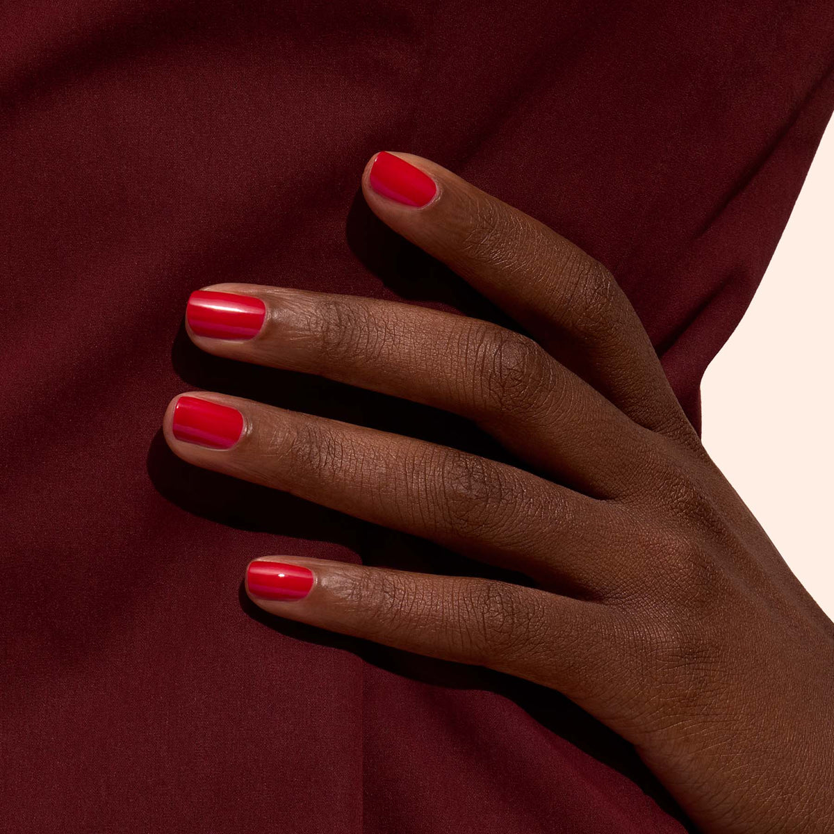 A hand with neatly manicured nails painted in Manucurist-test Poppy Red - Free rests on dark burgundy fabric, highlighting a striking color contrast.