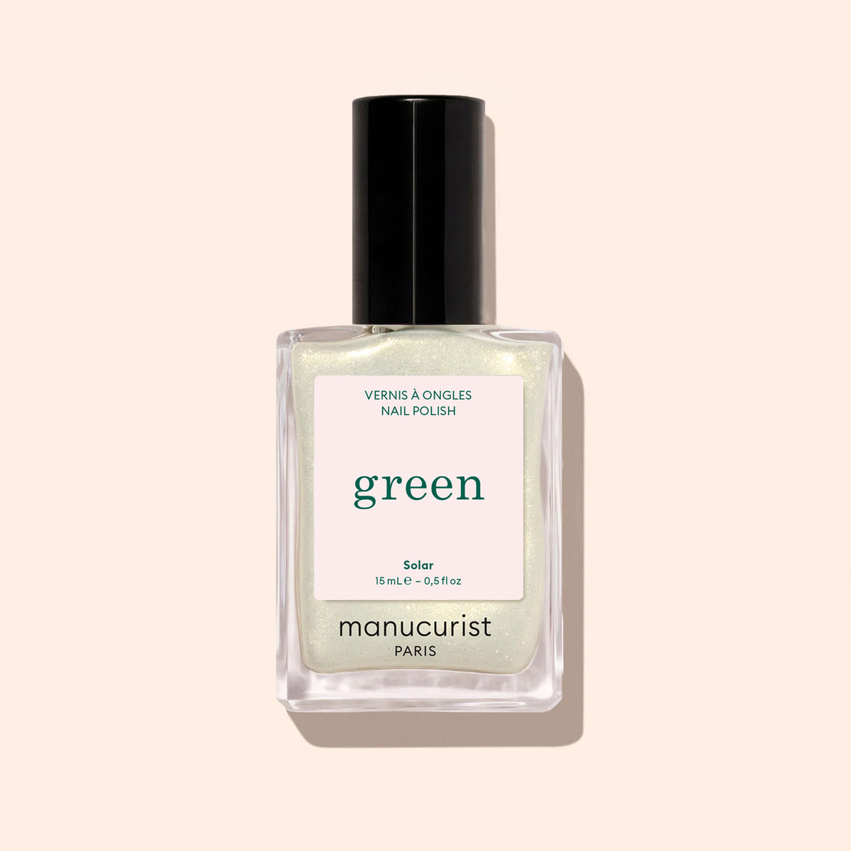 A bottle of Manucurist Solar nail polish, a light shimmery shade with golden highlights and a black cap, is displayed against a pale beige background.