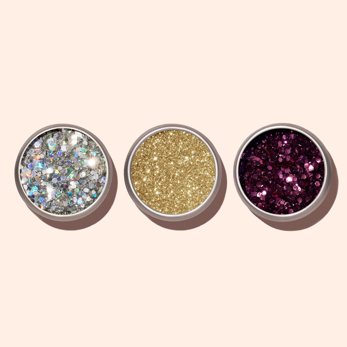 Three small round containers of Manucurist Holo biodegradable glitter are arranged side by side on a light beige background: silver holographic on the left, gold in the center, and dark pink-purple on the right.