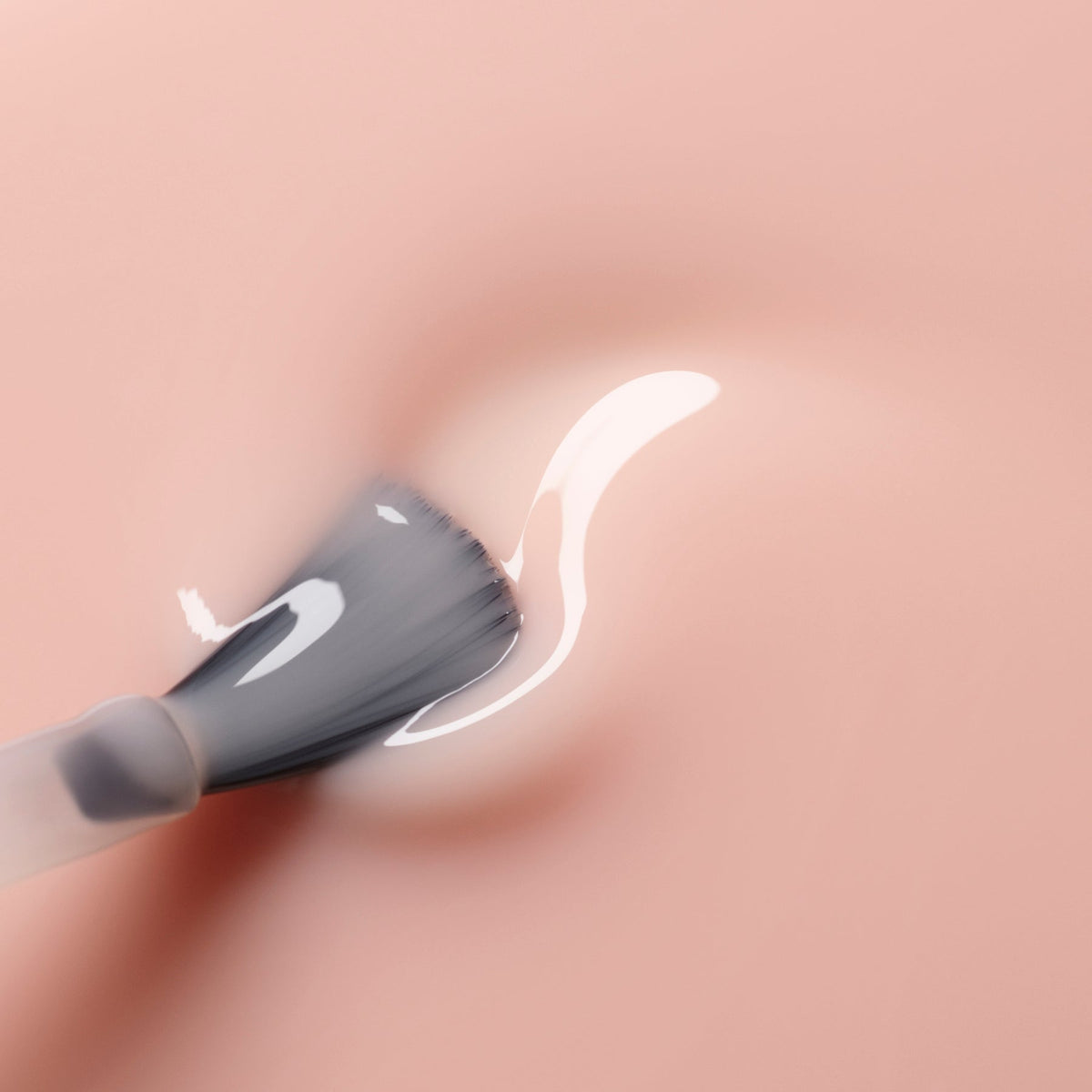 A close-up of a makeup brush applying Manucurist-test’s Pale Rose - Free, a creamy, sheer pastel pink nail polish, onto a smooth pink surface.