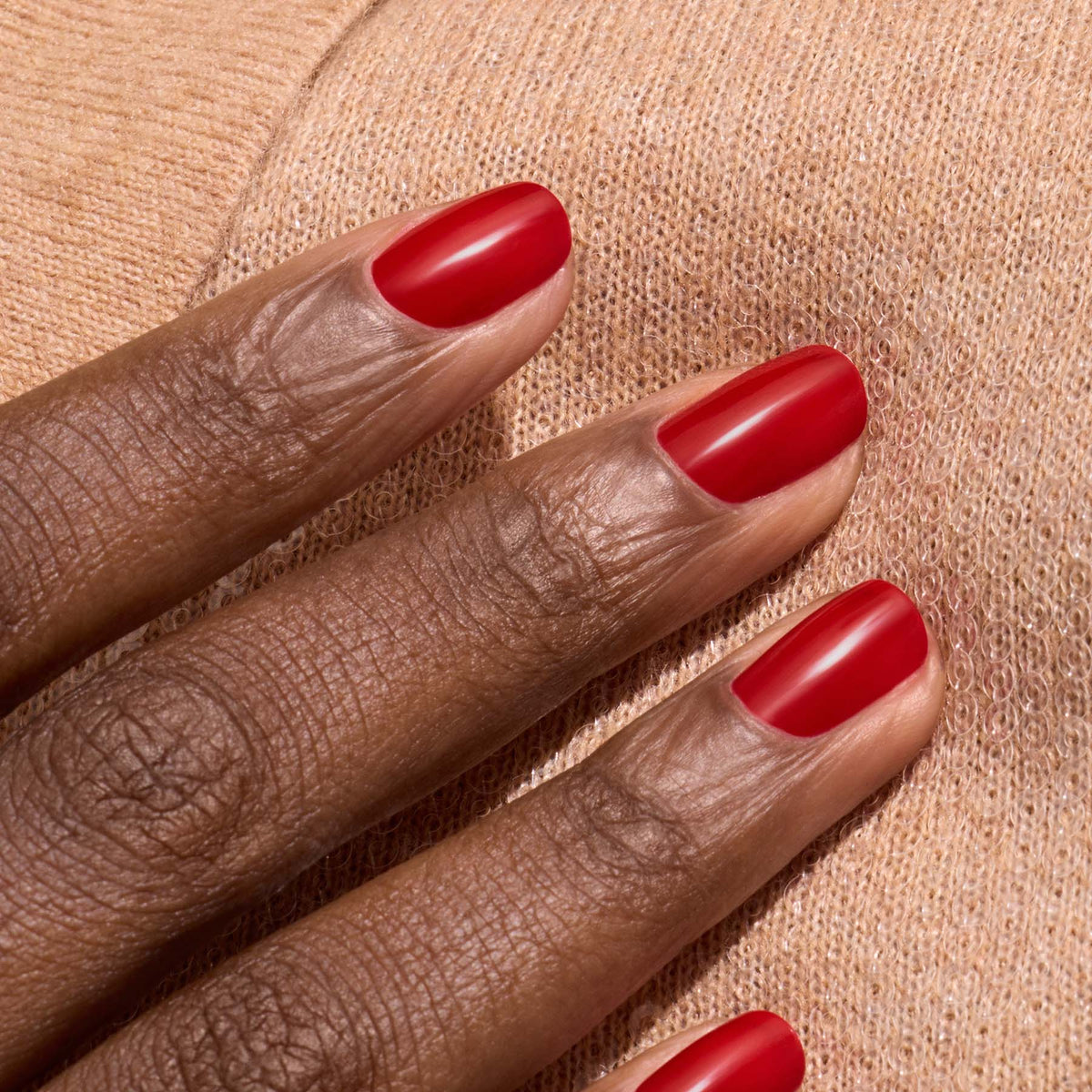 A close-up of a hand with dark skin and neatly manicured nails painted in Manucurist’s Red Cherry polish, resting on soft, light beige knit fabric.