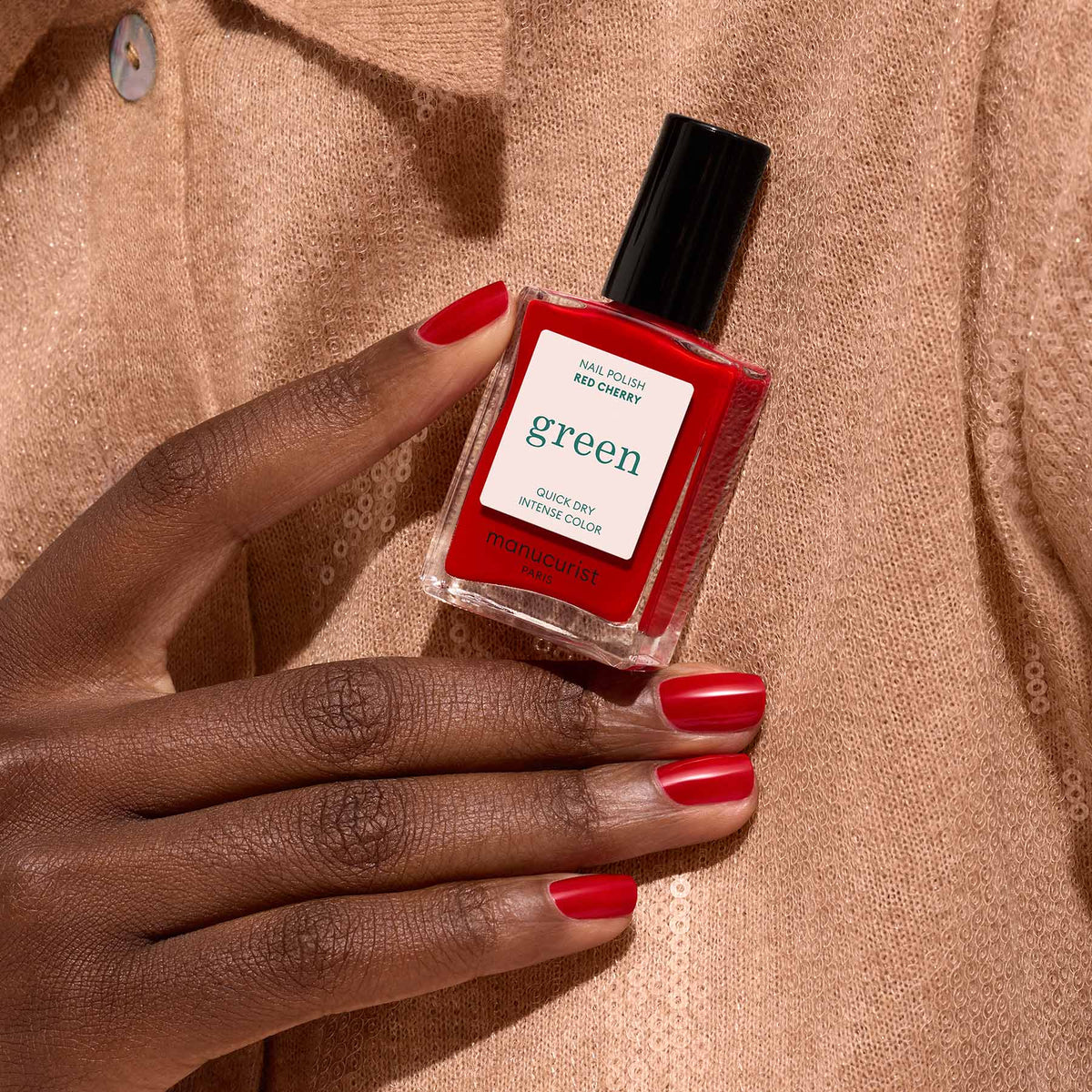 A hand with red painted nails holds a Manucurist Red Cherry nail polish bottle from the Green™ range against a beige, sequined fabric background.