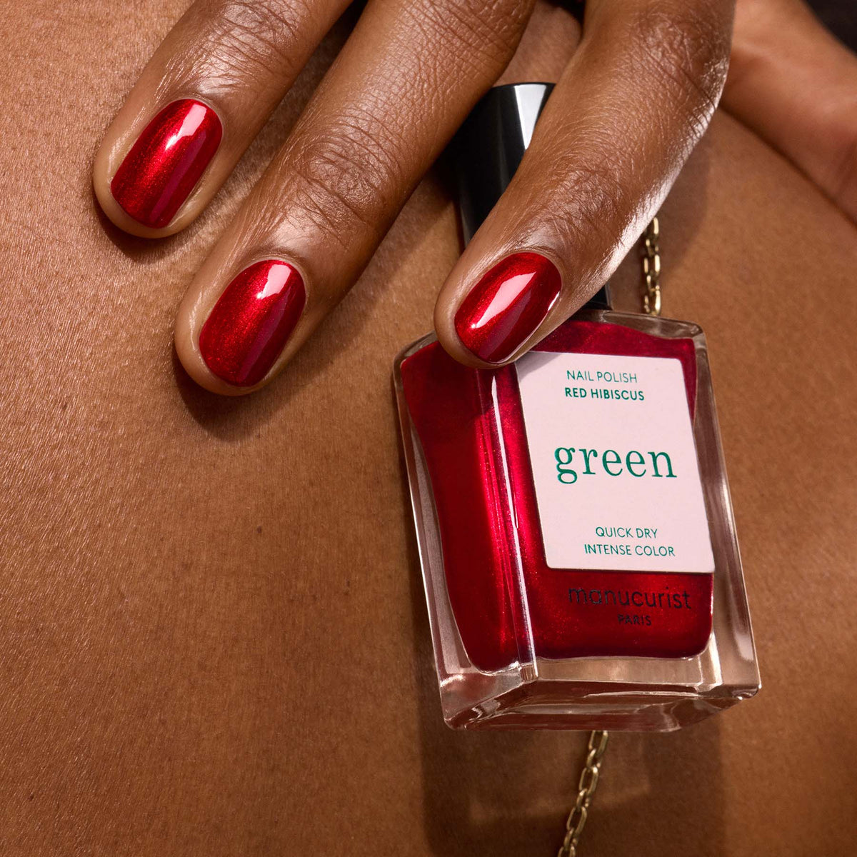 A hand with red polished nails holds a Manucurist Red Hibiscus nail polish bottle against a brown skin background.