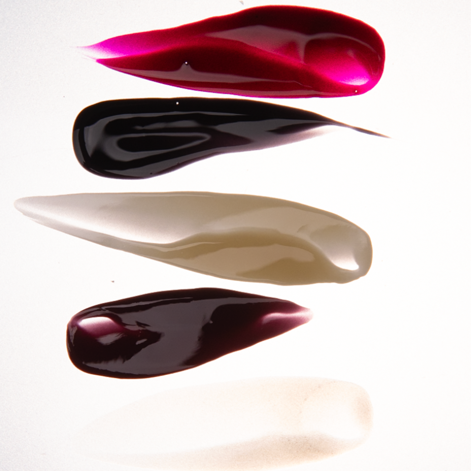 Five swipes of Manucurist’s Rainbow Collections nail polish in dark red, black, taupe, deep plum, and sheer white are arranged in sleek teardrops on a white background, highlighting curated modern manicure palettes.