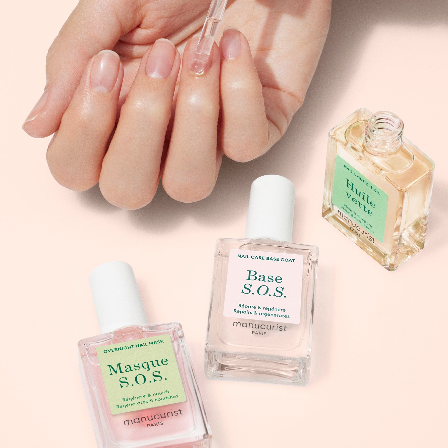 A hand applies oil to damaged nails with a dropper as three Manucurist Rescue Kit bottles are arranged on a light surface.