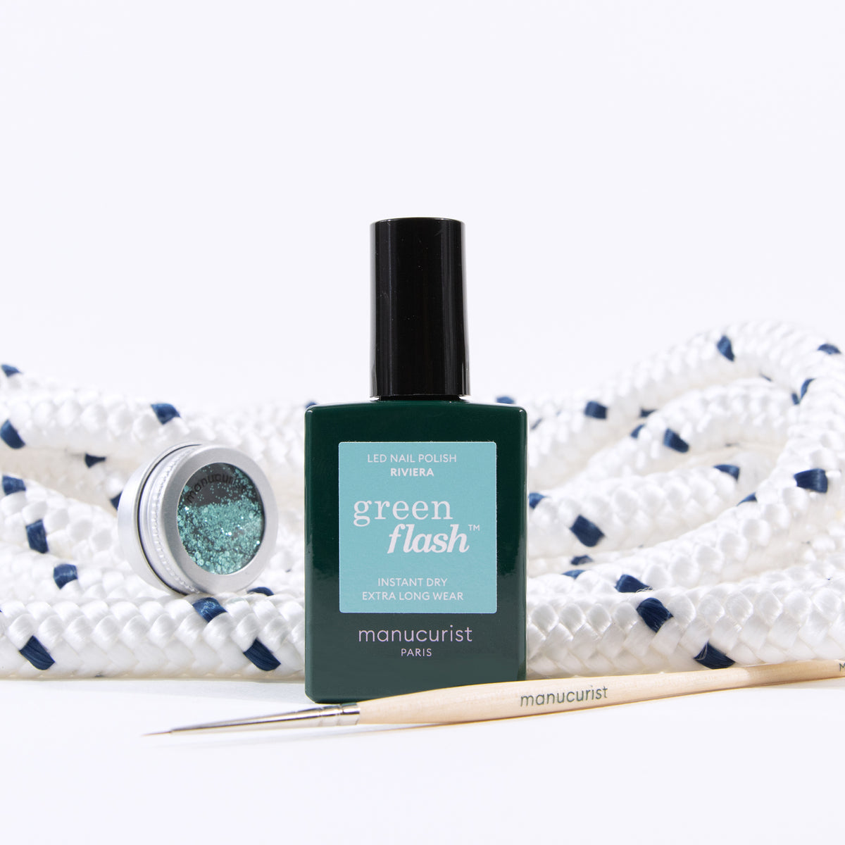A bottle of Manucurist Green Flash nail polish, a tin of Underwater glitter by Manucurist, and a wooden nail art brush are arranged before a coiled white rope with blue accents.