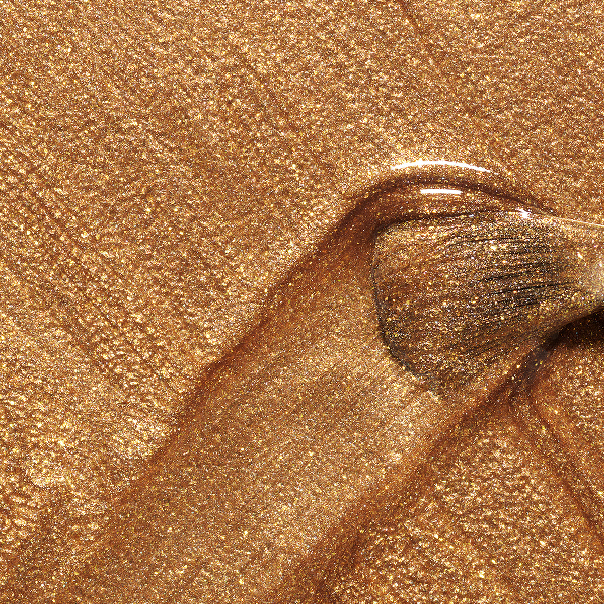 A close-up of a makeup brush spreading shimmery gold liquid foundation on a glittery surface, evoking the radiant glow of Shimmer by Manucurist.