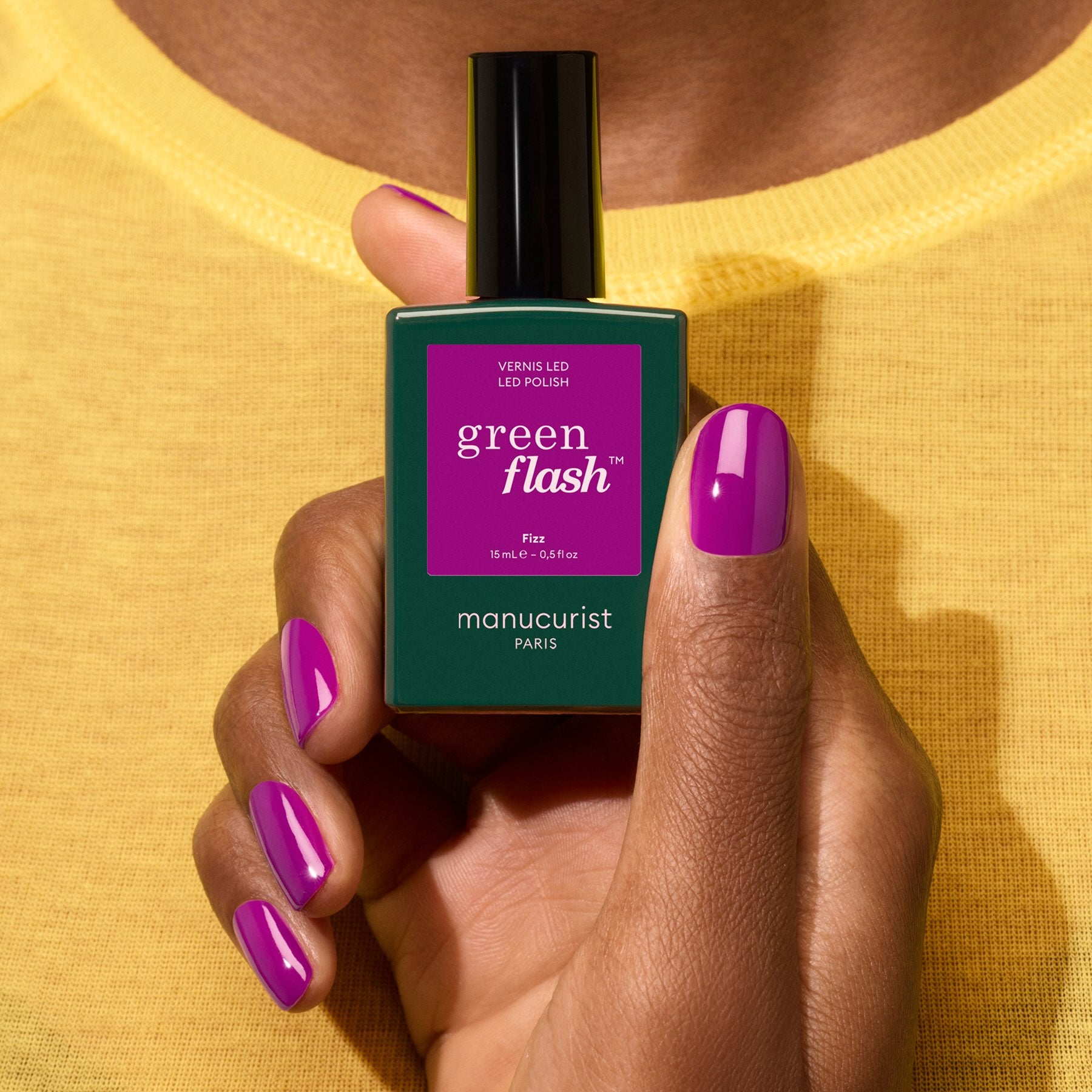 A hand with shiny purple nail polish with magenta undertones holds a green bottle labeled Fizz by Manucurist, set against a yellow shirt background.