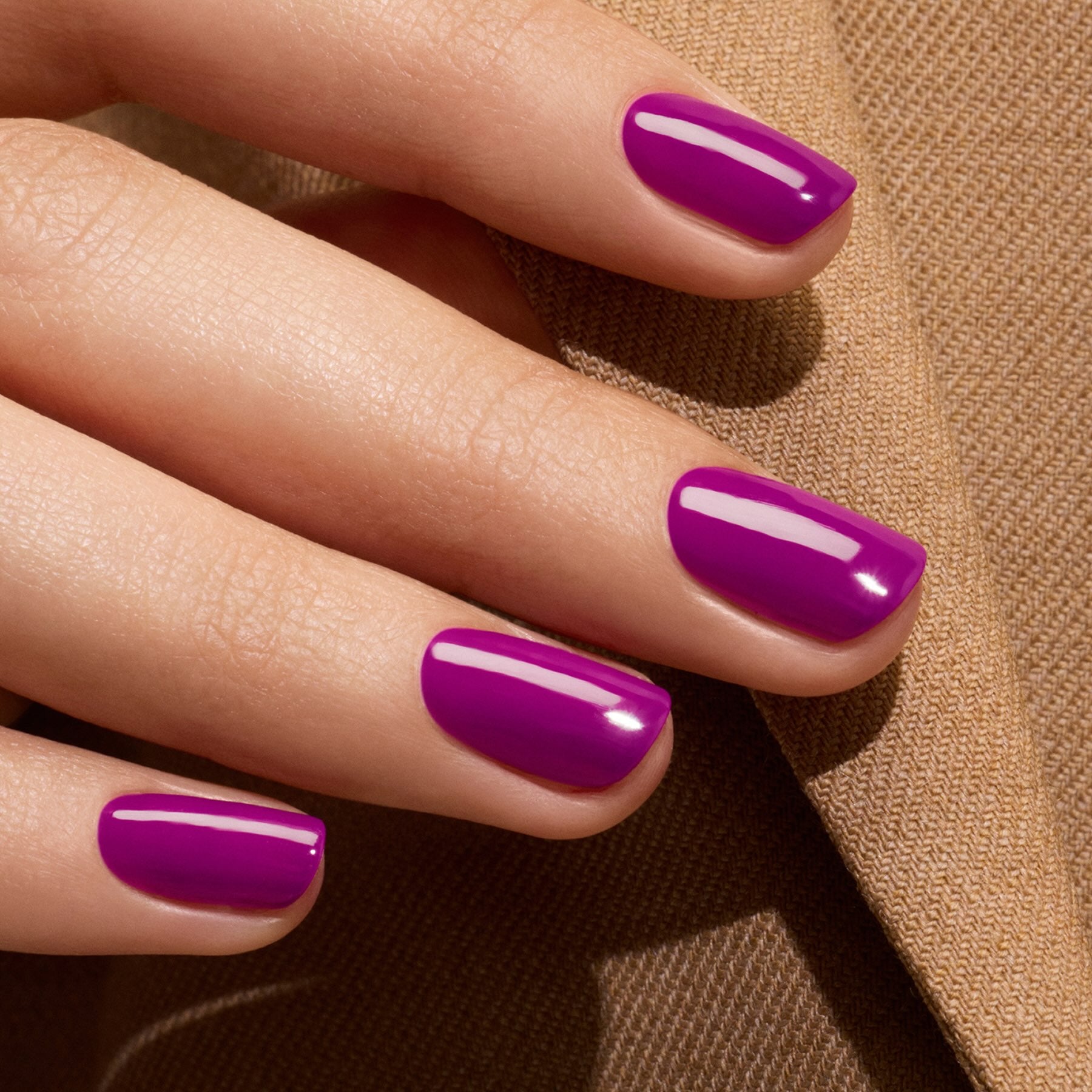A close-up of a hand with neatly manicured nails painted in Manucurist’s Fizz, a glossy, vibrant violet gel polish with magenta undertones, resting on beige fabric.