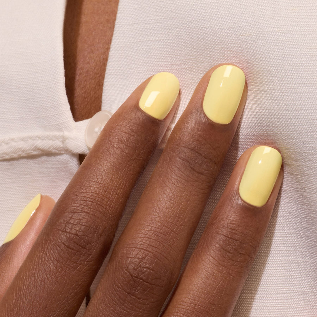 A close-up of a hand with dark skin shows neatly manicured nails painted in glossy pastel yellow Mimosa nail polish by Manucurist, resting on white fabric with a button detail.