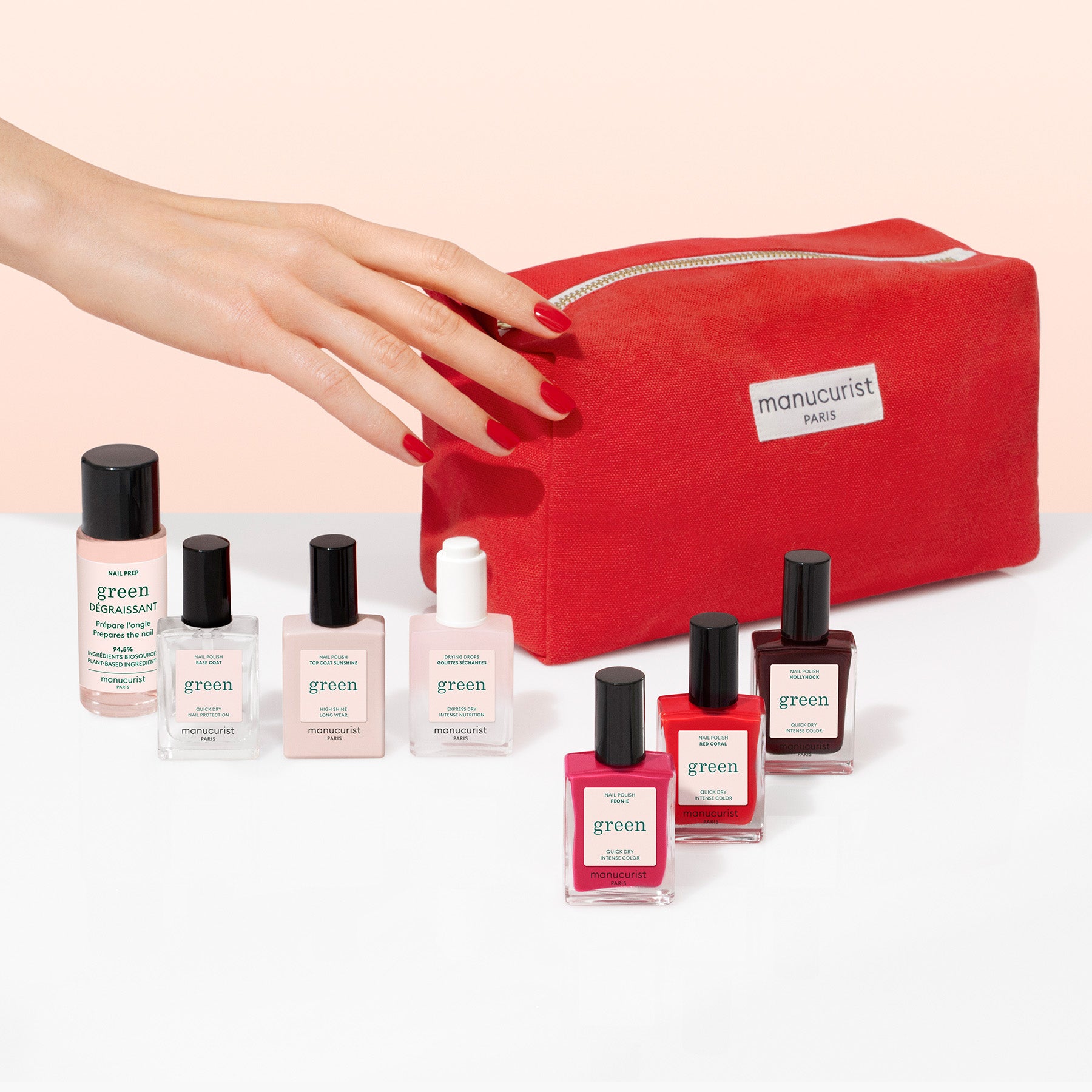 A hand with red nail polish reaches for a red Manucurist Starter Kit bag, surrounded by seven plant-based nail polishes and nail care products in assorted shades against a light gradient background.