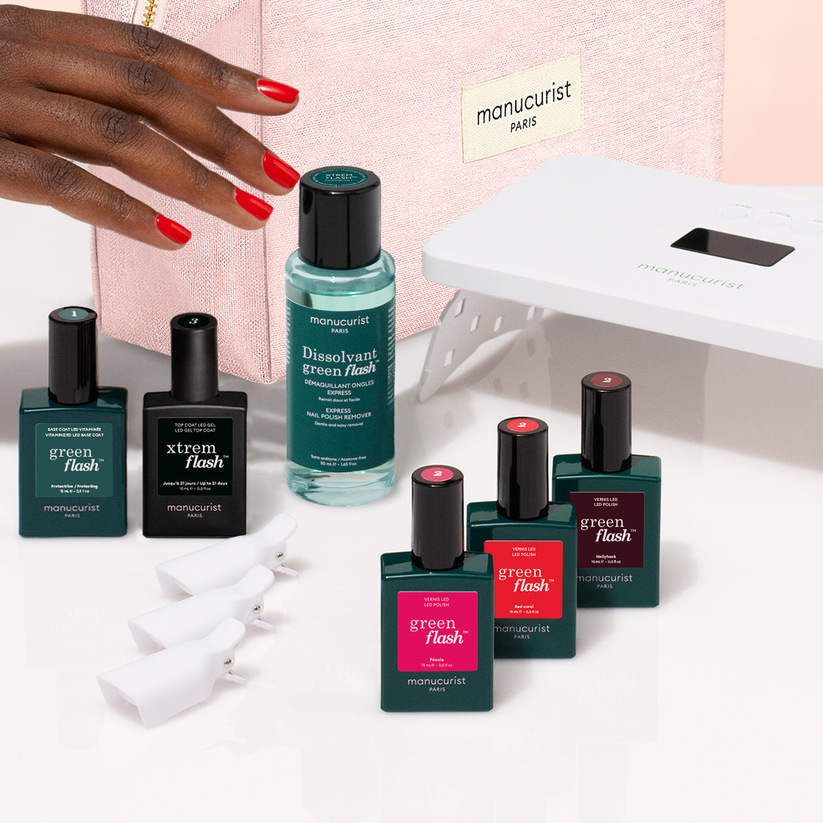 A hand holds a pink Manucurist Starter Kit manicure bag near nail polishes, remover, clips, and an LED lamp from the Manucurist Starter Kit, all displayed on a white surface.