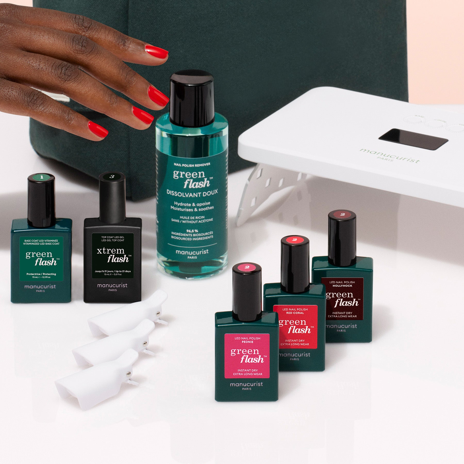 A hand with red nail polish reaches for the Manucurist Starter Kit, which includes a gel polish kit, remover, clips, and a white LED lamp on a clean white surface.