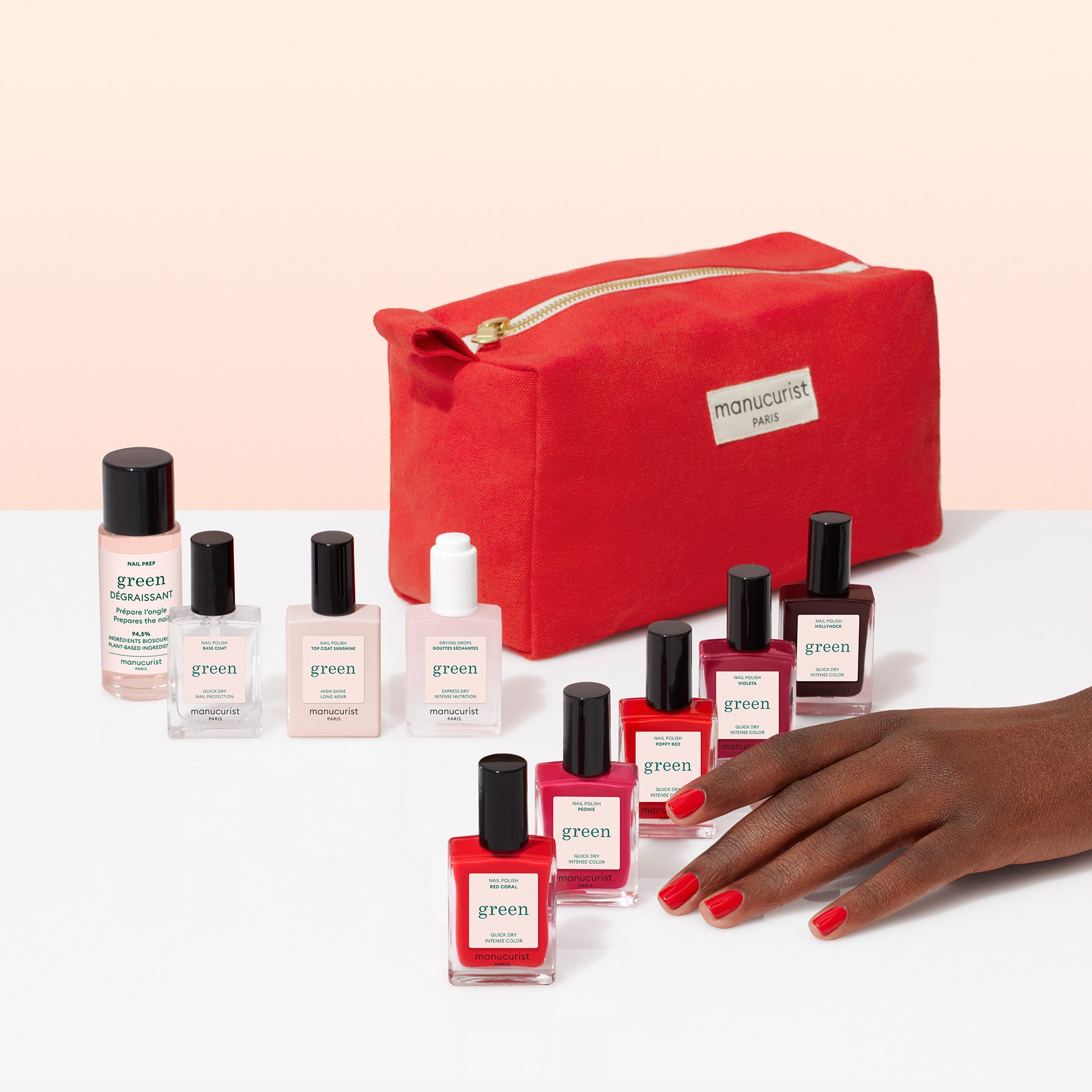 A hand with red-painted nails is shown beside several Manucurist Super Starter Kit plant-based nail polishes and manicure products, all arranged in front of a red zippered pouch on a white surface with a peach background.