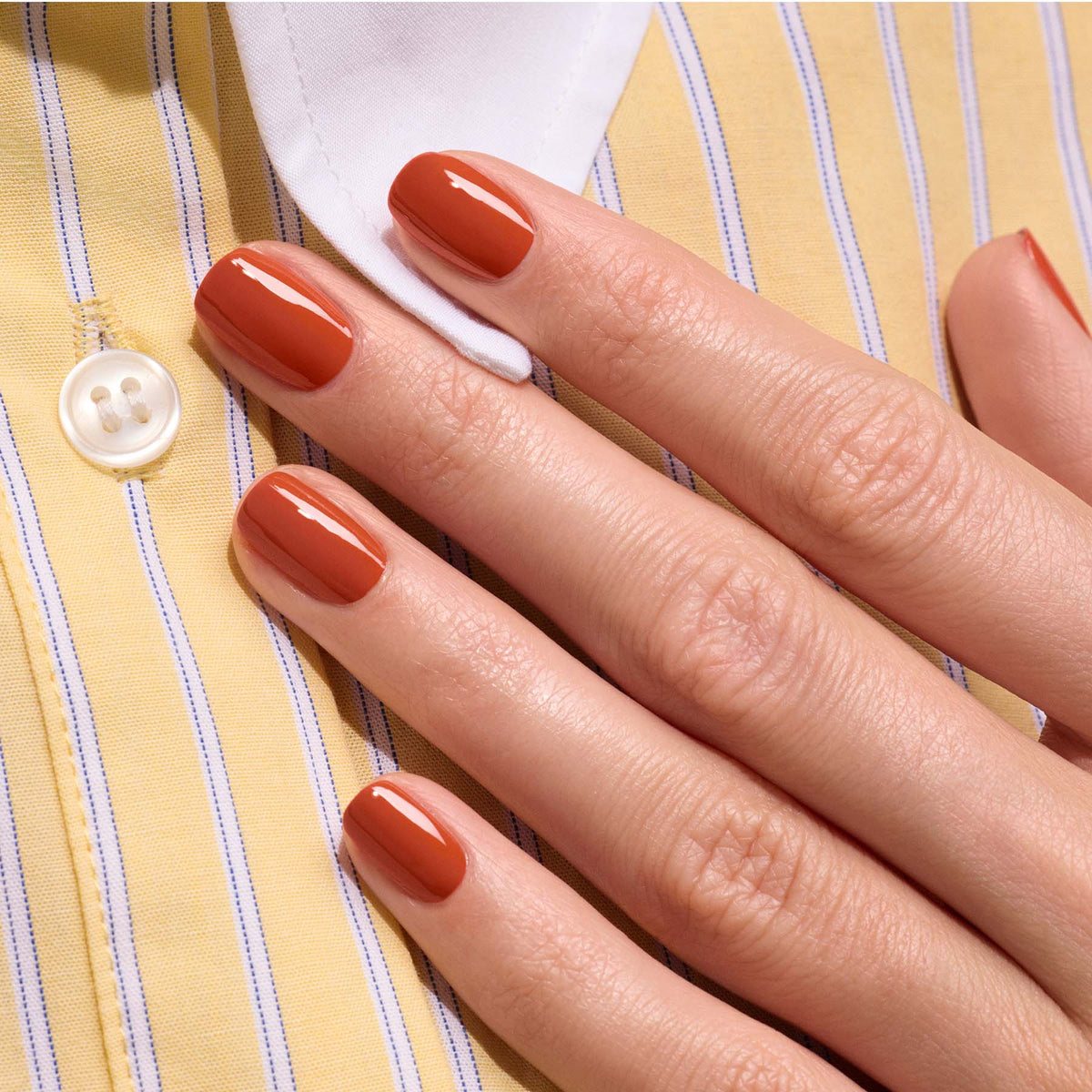 A hand with neatly manicured nails painted in Manucurist’s Green™ Terracotta, a glossy scorched red earth shade, rests on a yellow and white striped shirt with a white button.