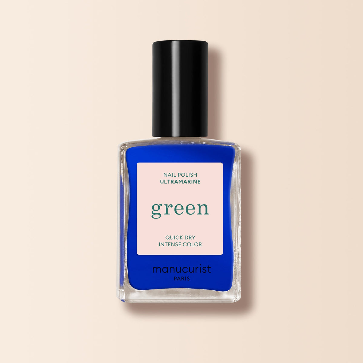 A bottle of Manucurist Ultramarine nail polish in bold blue with a black cap, labeled "Nail Polish Ultramarine, Green, Quick Dry Intense Color," is set against a soft beige background.