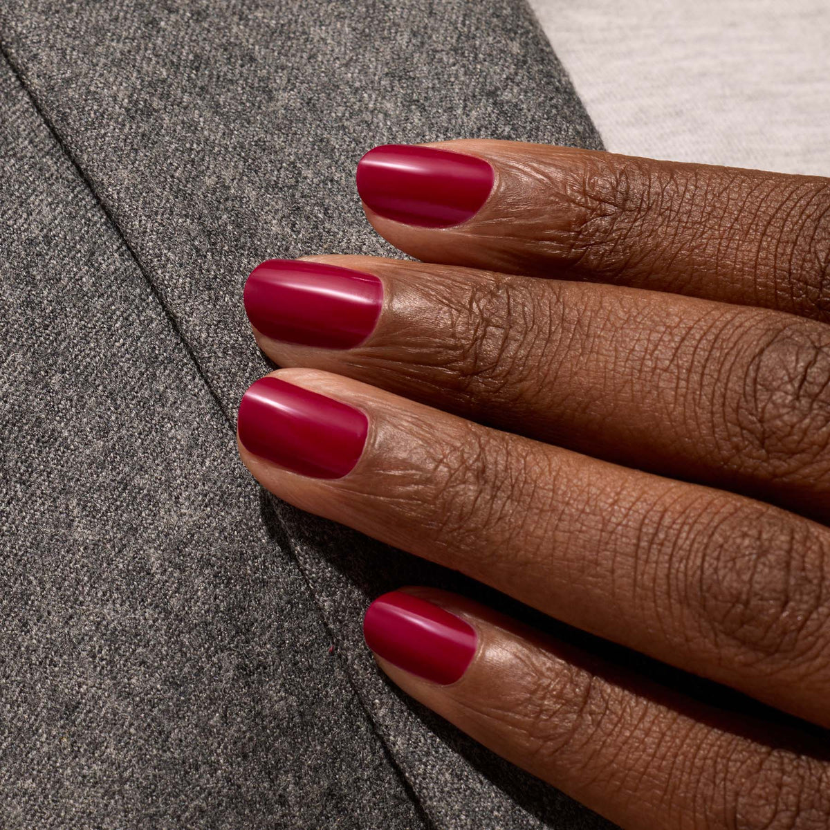 A hand with medium-dark skin tone displays neatly manicured nails painted in Manucurist’s Violeta, a rich burgundy purple shade, resting on a gray fabric surface.