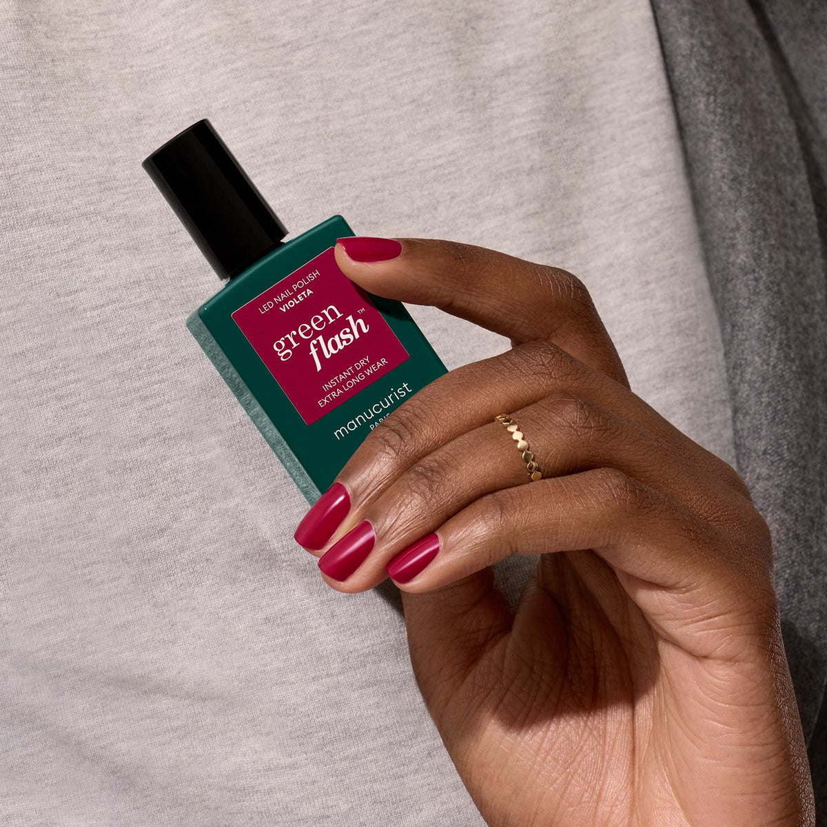 A hand with neatly manicured violet nails holds a bottle of Manucurist Violeta gel polish against a light gray fabric background, with a delicate gold ring on one finger.