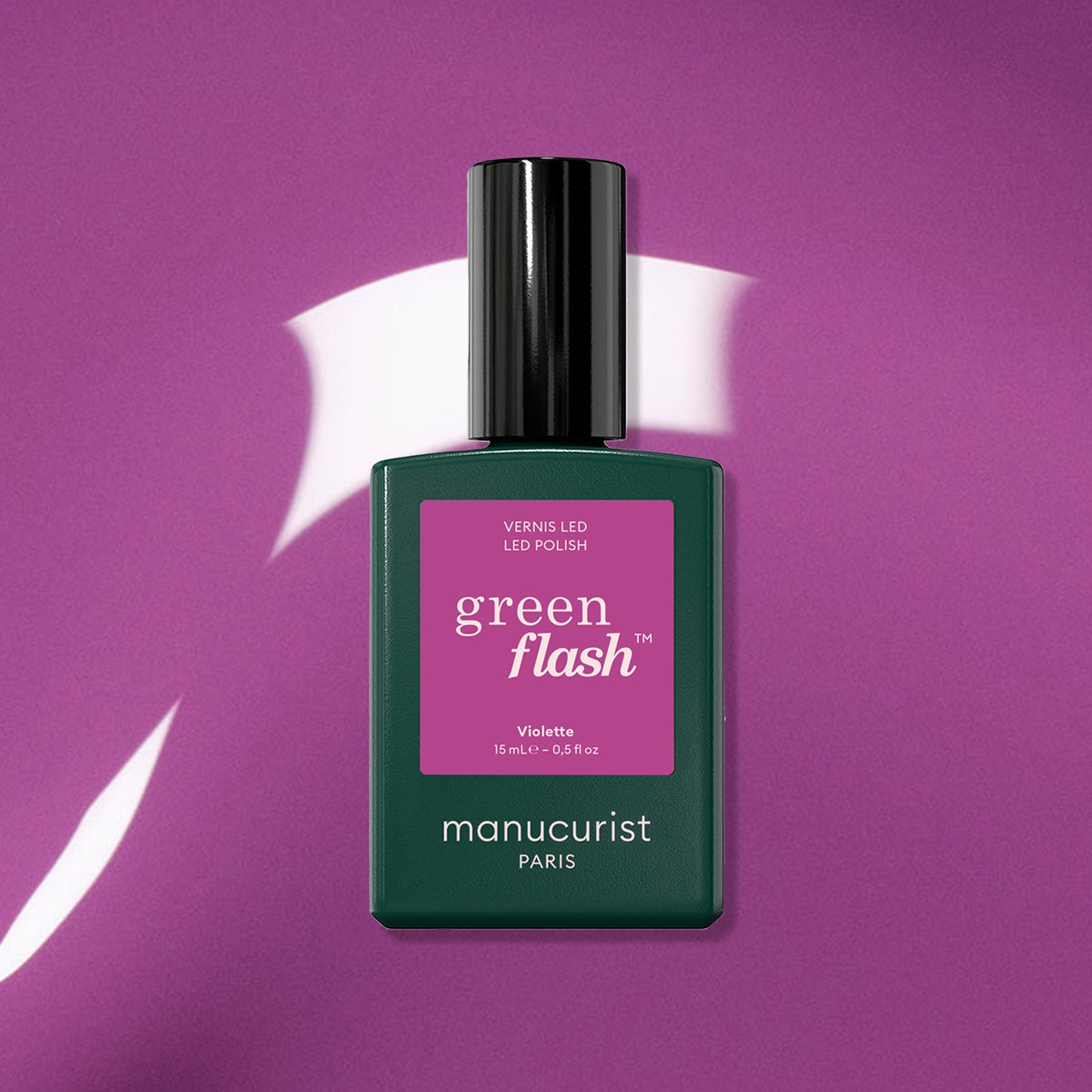 A bottle of Manucurist’s Violette Green Flash gel polish, in a dark green bottle with a rectangular pink label and sleek black cap, rests on a retro purple surface.