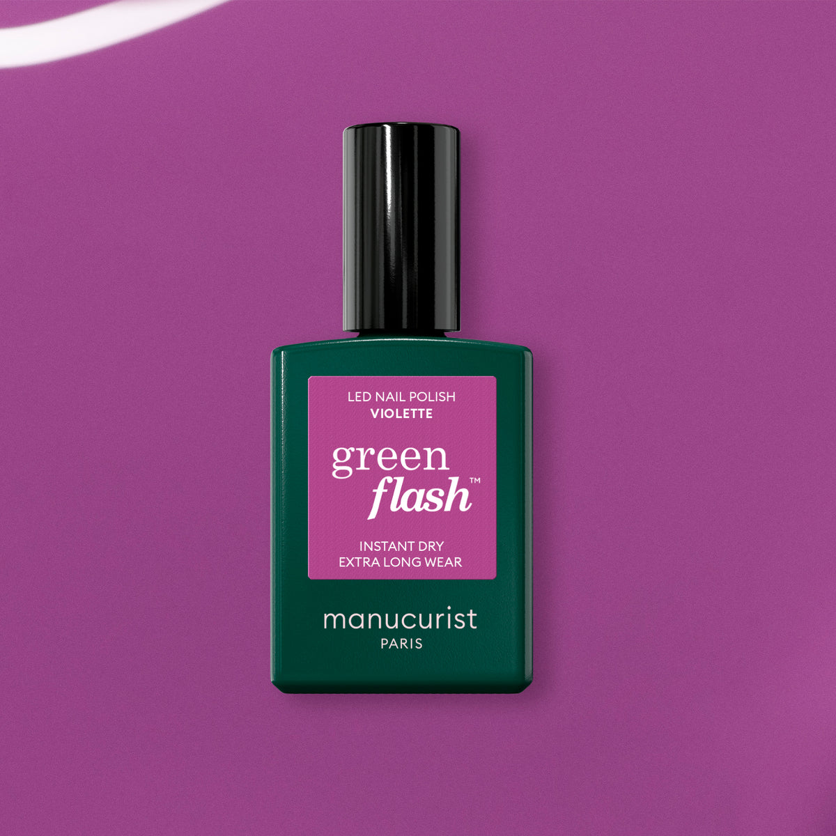A bottle of Manucurist’s Violette Green Flash gel polish, in a dark green bottle with a rectangular pink label and sleek black cap, rests on a retro purple surface.